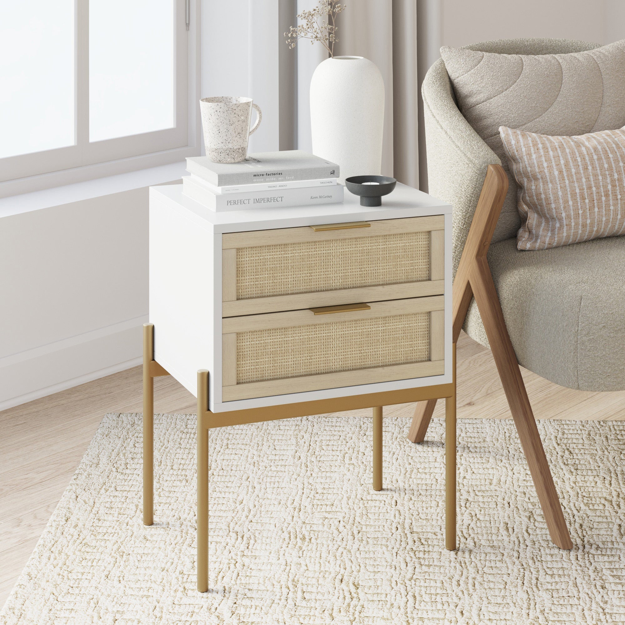 The Nathan James Wood & Rattan 2-Drawer Nightstand in white, featuring cane drawers and gold legs, stands by a light armchair. On top sit books, a speckled mug, a white vase, and a small black dish as sunlight streams through the window.