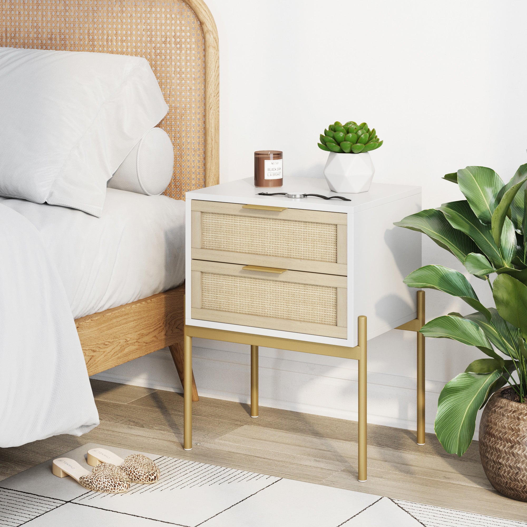 A modern bedside table with two drawers and gold legs holds a potted succulent, a brown candle, and a book, next to a bed and a leafy green plant on a light wood floor.