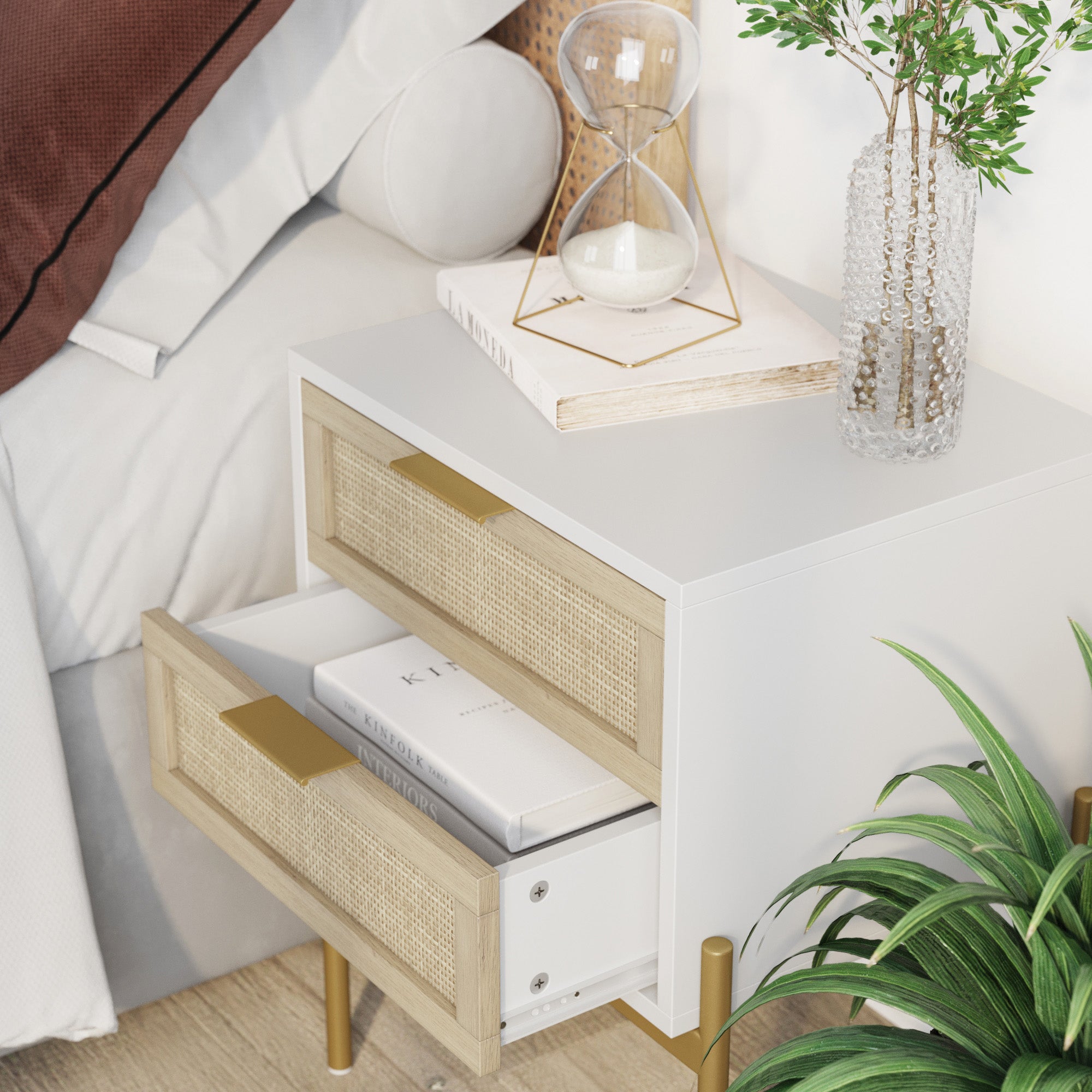 A modern white nightstand with wooden drawers sits beside a bed. One drawer is open, showing books inside. On top are an hourglass, a stack of books, and a textured glass vase with greenery. A leafy plant is nearby.