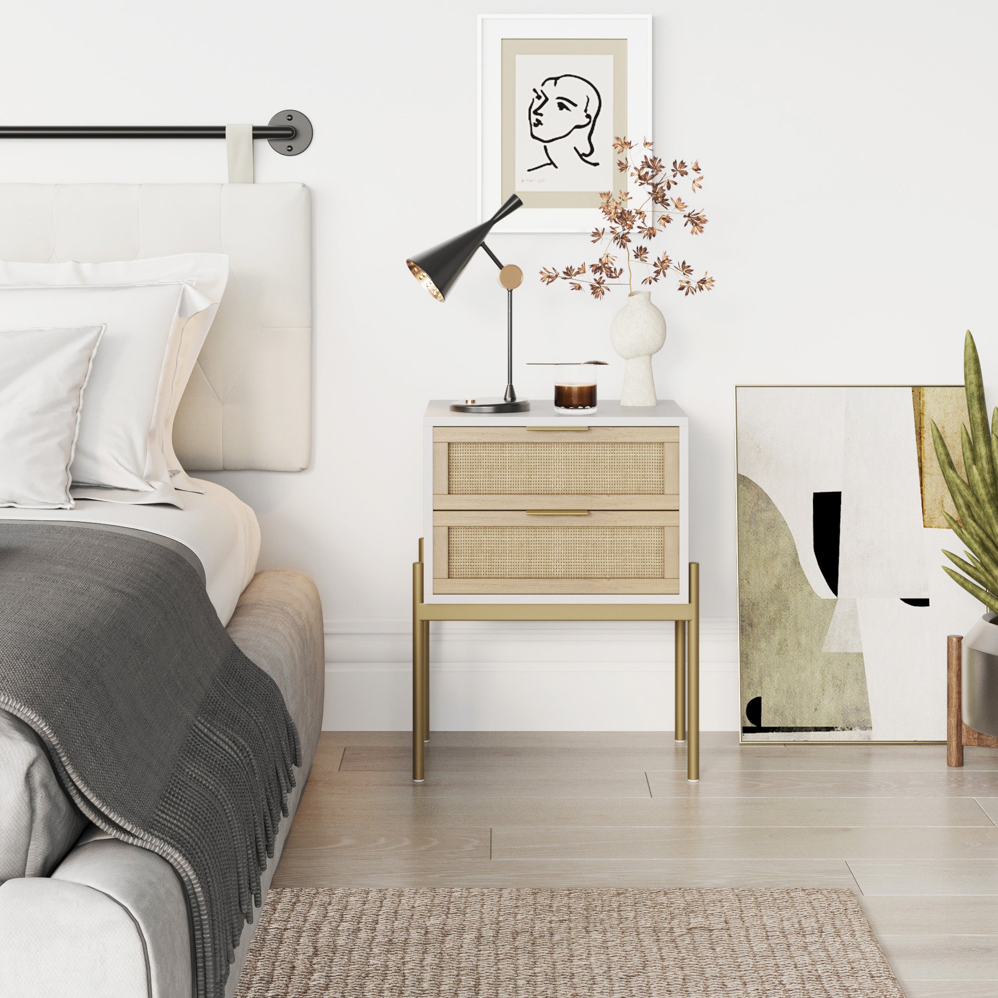 A modern bedroom with a white bed, gray blanket, and a light wood nightstand holding a black lamp, vase with dried branches, and small decorations. Abstract artwork is propped against the white wall next to a tall green plant.