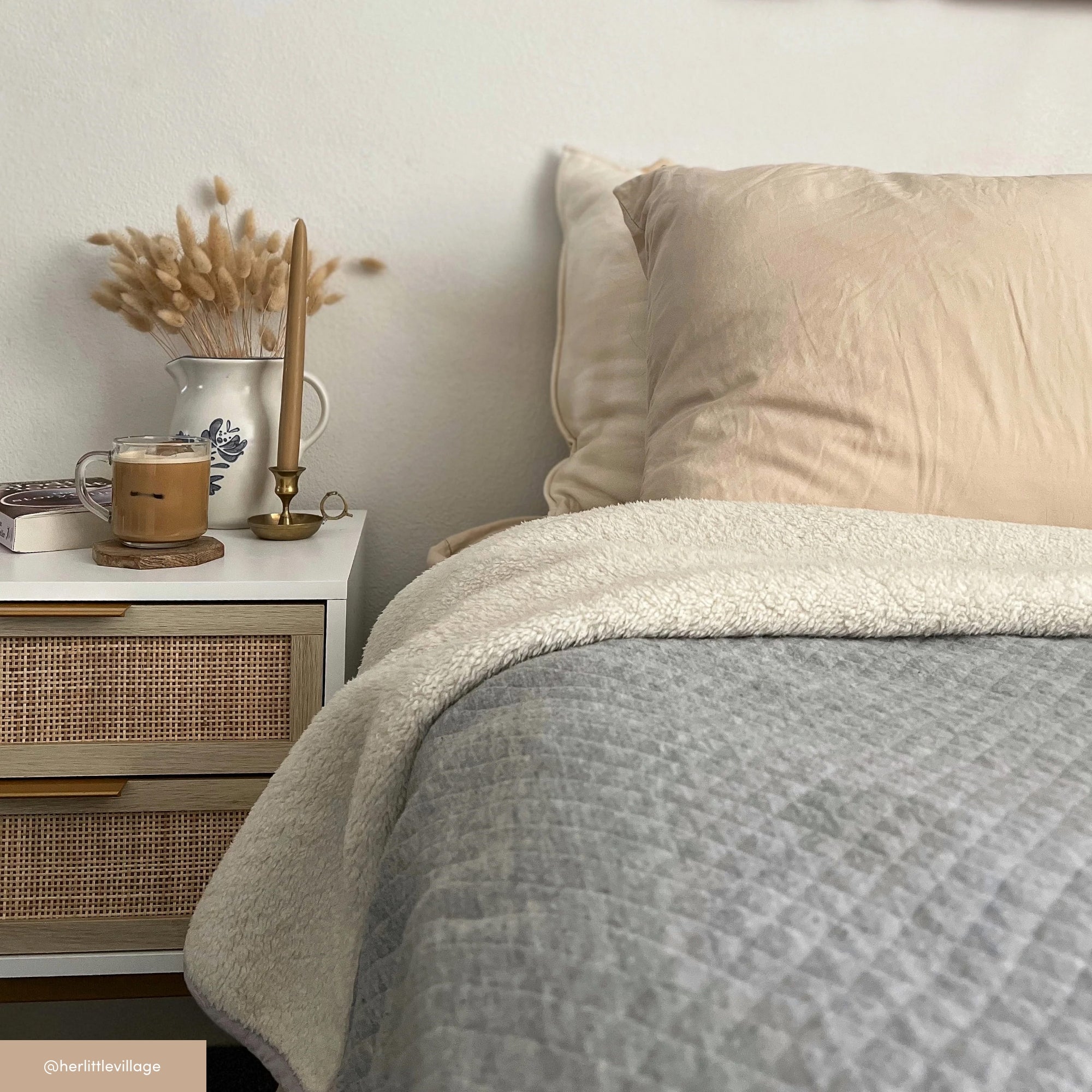 A cozy bedroom scene with a bed made up in beige and gray bedding, next to a nightstand holding a ceramic jug with dried flowers, a lit candle, and a glass mug of coffee.