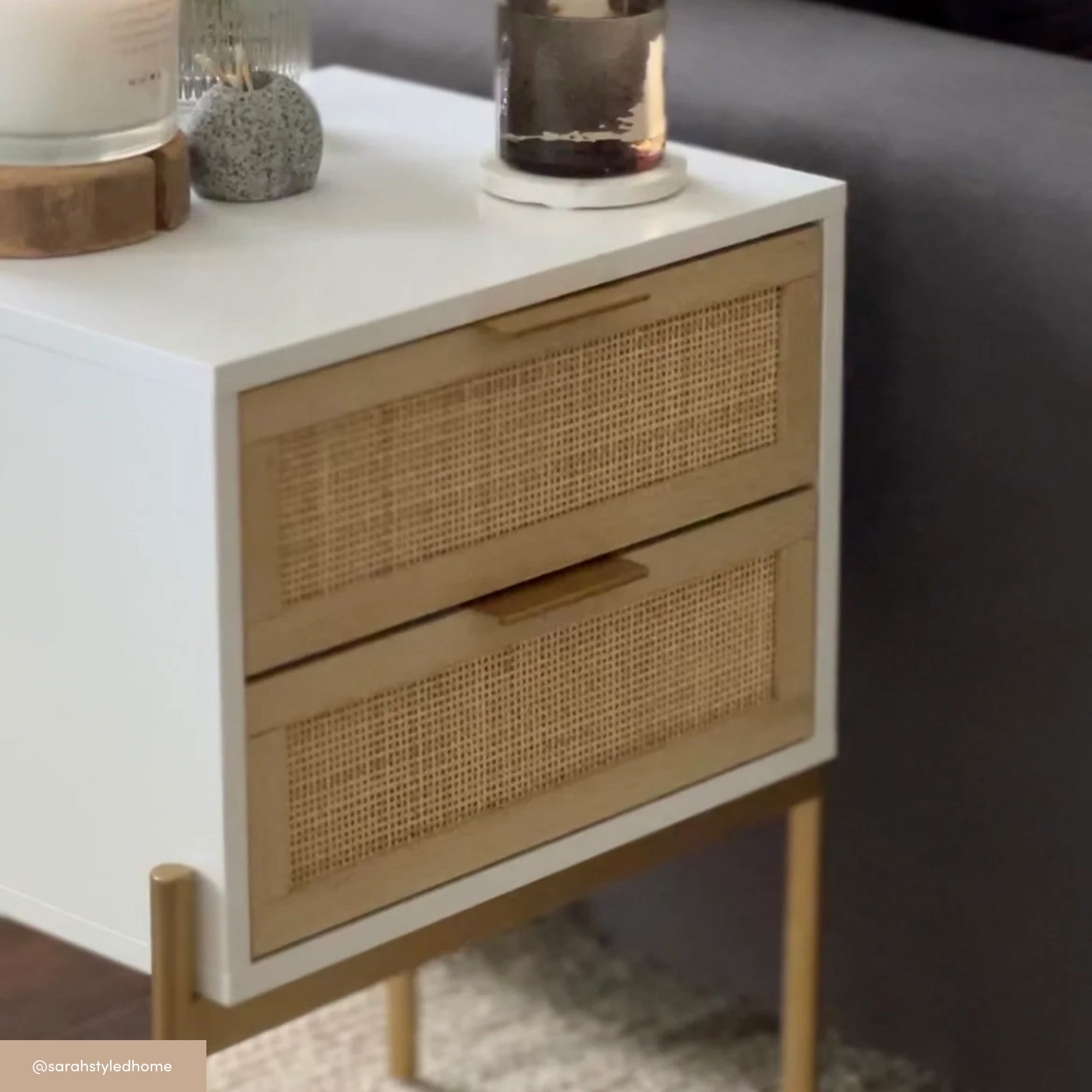 A modern white and gold nightstand with two cane-front drawers beside a gray sofa. The top holds a candle, small decorative items, and a glass container.