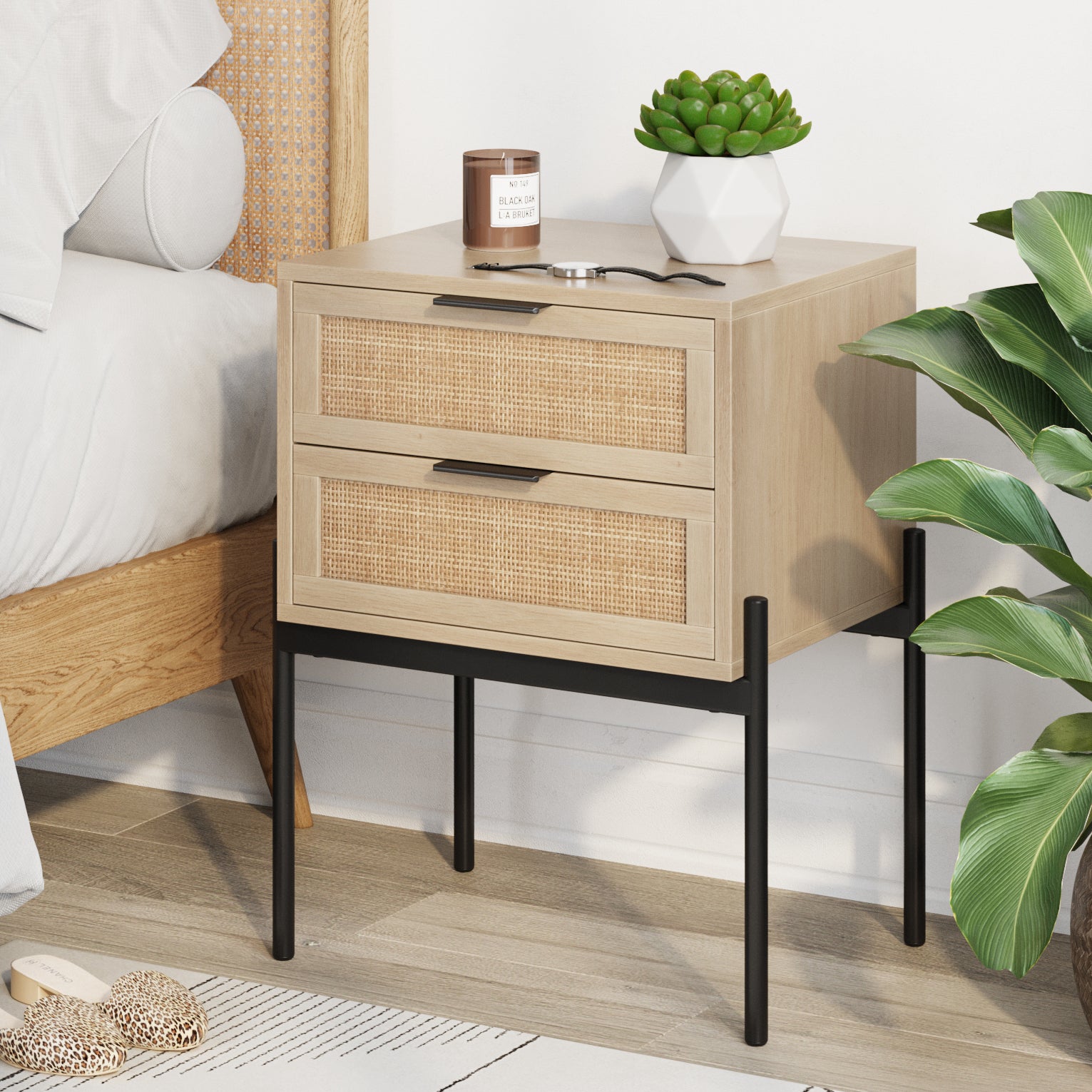 A modern bedside table with two drawers featuring wicker fronts, black legs, and a light wood finish. On top are a succulent plant, a candle, and glasses. Nearby are a bed, a leafy plant, and slippers on the floor.