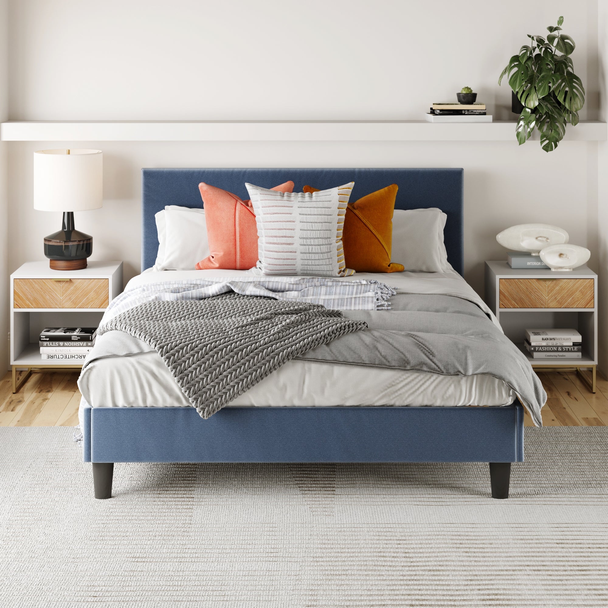 A modern bedroom features a blue upholstered bed, gray and white layered bedding with colorful pillows, and Nathan James Wood Herringbone Nightstands (Set of 2) in natural finish, topped with a lamp, books, and decor. A potted plant sits on the wall shelf above.