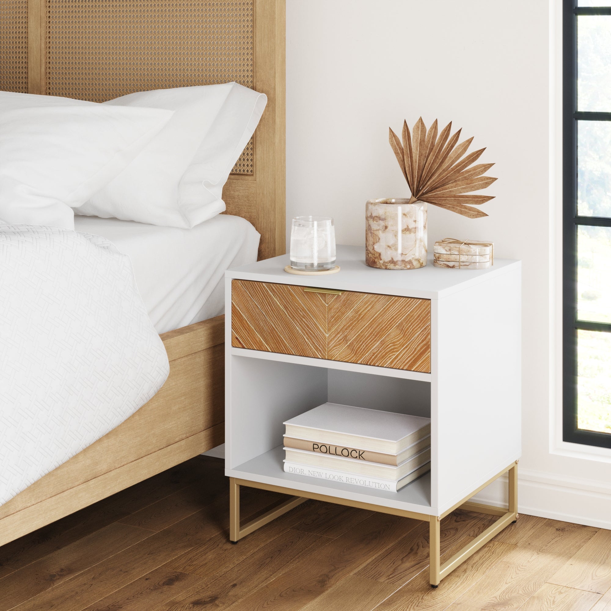 The Nathan James Wood Cabinet Herringbone Nightstand in Natural Wash, with a drawer and open shelf, holds a glass of water, vase with dried palm leaves, coasters, and books beside a bed with white bedding near a black-trimmed window.