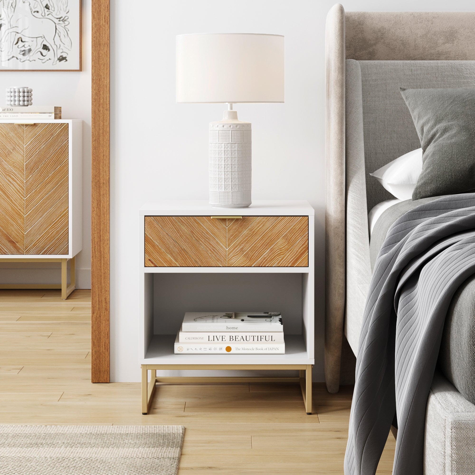 A modern bedside table with a wood and white finish features a lamp, books, and decor. The table is next to a gray upholstered bed with a gray blanket, on light wood flooring in a bright, stylish bedroom.