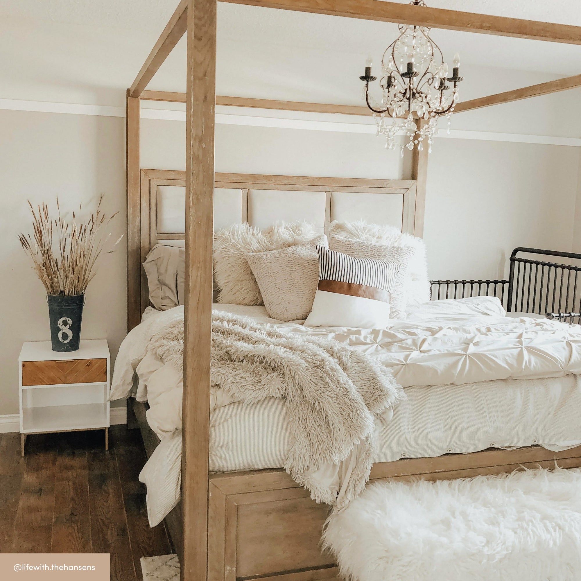 A cozy bedroom with a wooden four-poster bed, fluffy white bedding, assorted pillows, a chandelier, a white nightstand with a vase of dried grass, and wooden floors. The decor is soft and neutral.