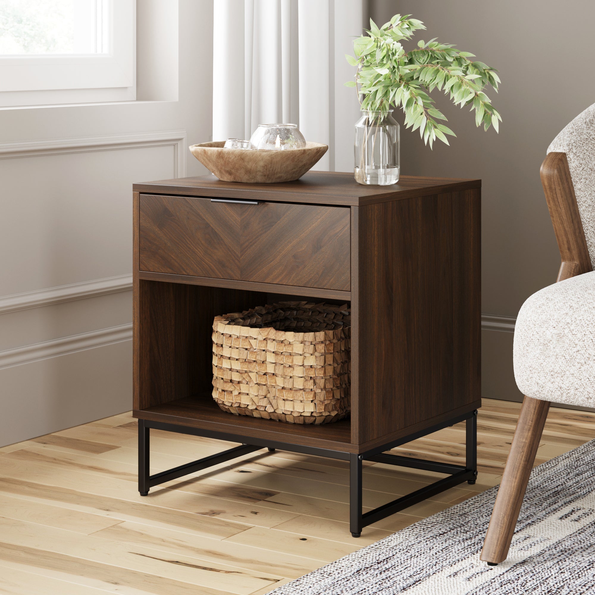 The Nathan James Wood Cabinet Herringbone Nightstand in walnut features a drawer, open shelf with a woven basket, black metal legs, and is styled with a bowl, glass, and leafy plant on top beside a chair and window.