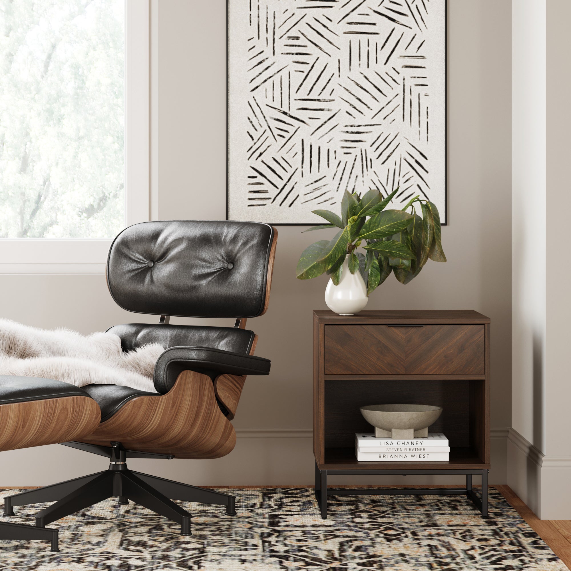 A modern lounge chair with wood and black leather sits by a window. Next to it is a wooden side table with a potted plant, books, and a bowl, beneath abstract black-and-white wall art.