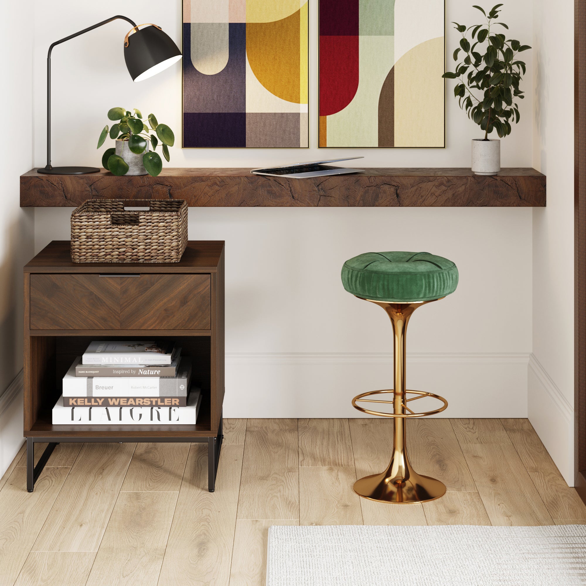 A modern workspace with a floating wooden desk, a green velvet stool with a gold base, a table lamp, potted plants, a woven basket, and a wooden side table with stacked magazines on a light wood floor.