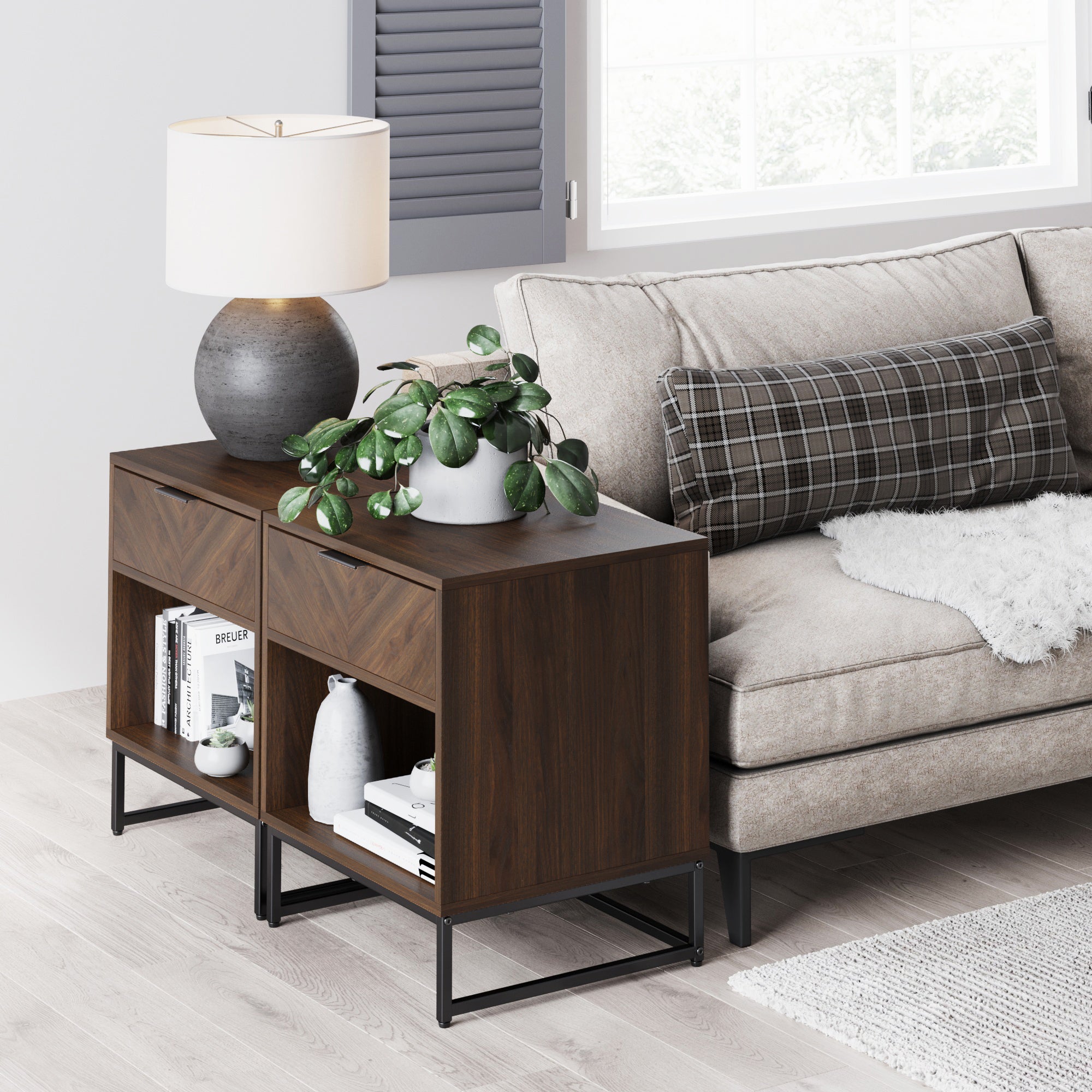A modern living room with a dark wood side table beside a beige sofa. The table holds a gray lamp, a green potted plant, books, and decorative objects. A window lets in natural light.