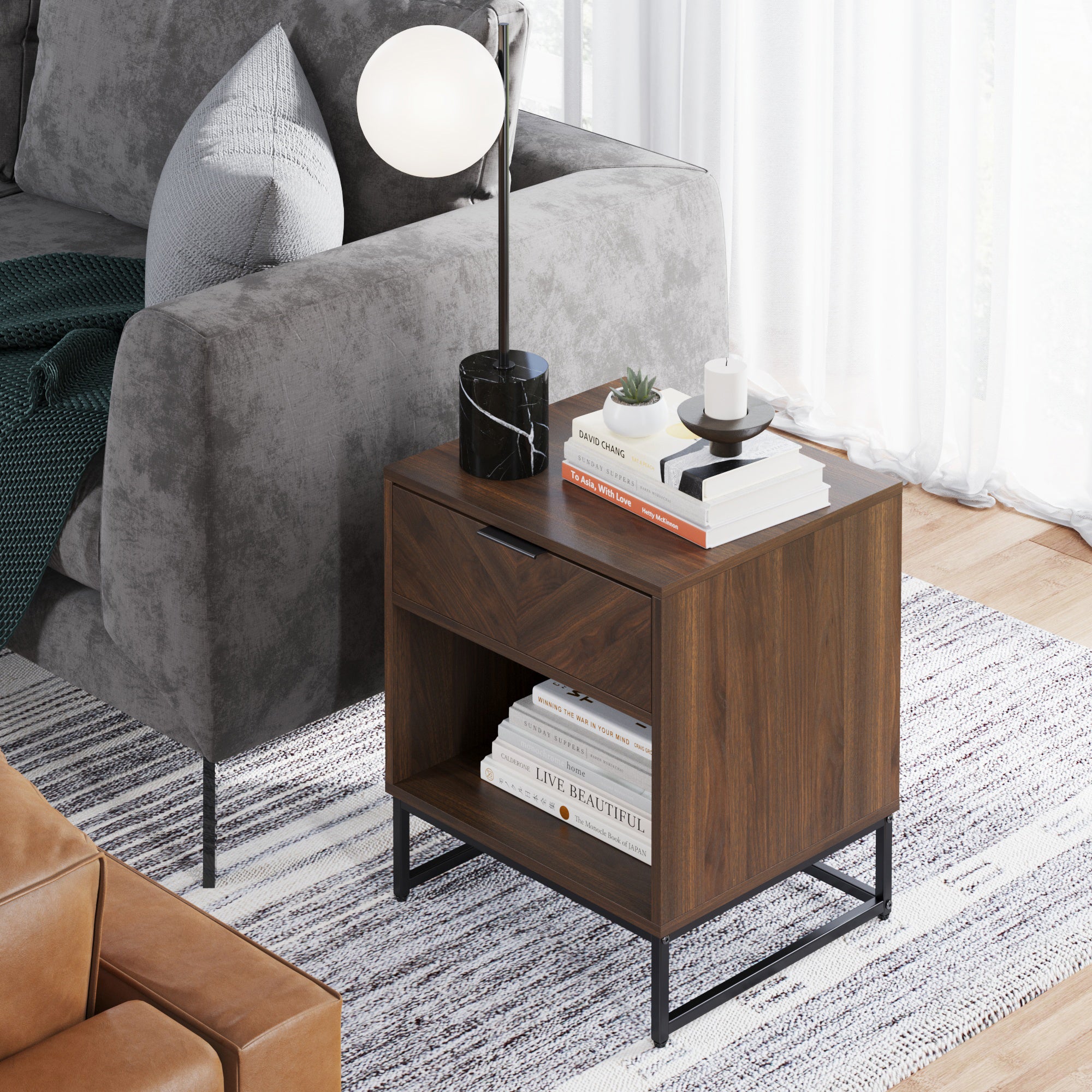 A modern wooden side table with a drawer and shelf stands beside a gray sofa. On top are a lamp, small plant, and candle; books are stacked on the shelf below. Sunlight streams through white curtains onto the rug.