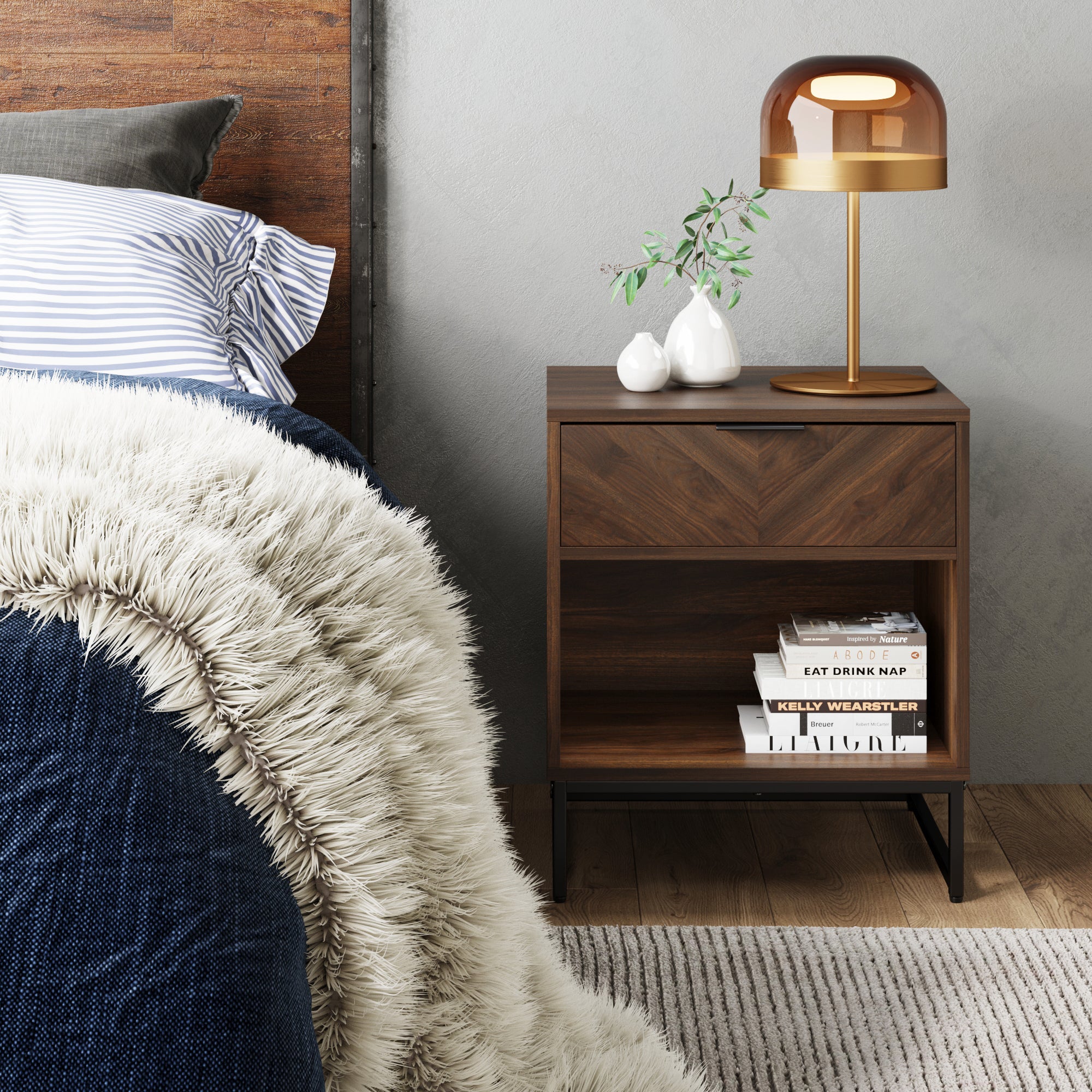 A modern bedroom scene with a dark wood nightstand holding a gold lamp, two white vases with green branches, and stacked books. The bedside has a navy blanket and a striped pillow, with a textured fur throw.