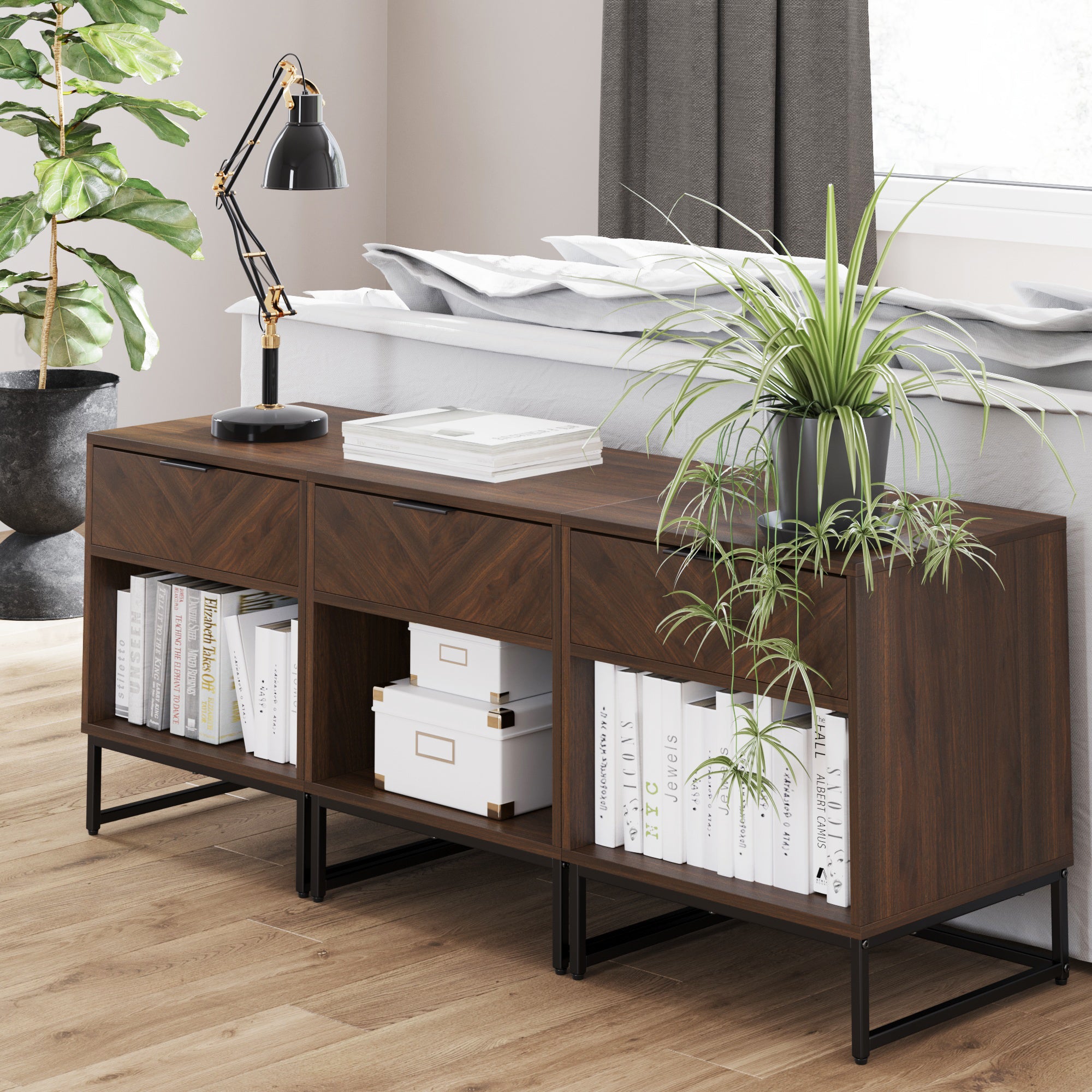 A modern wooden sideboard with open shelves, books, storage boxes, a table lamp, and a potted plant sits on a light wood floor next to a sofa and a tall leafy plant by a window with gray curtains.