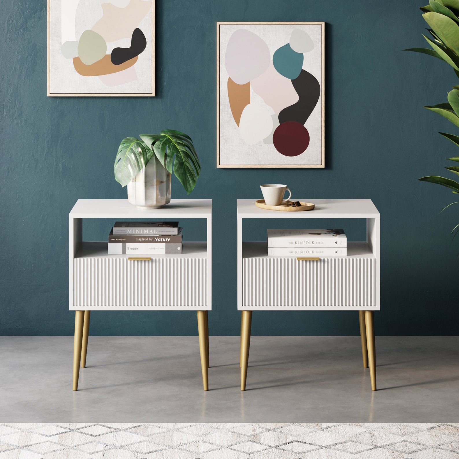 Two modern white nightstands with gold legs stand against a dark teal wall. Each holds books and decorative items. Abstract art hangs above them, and a plant is visible on the right side.
