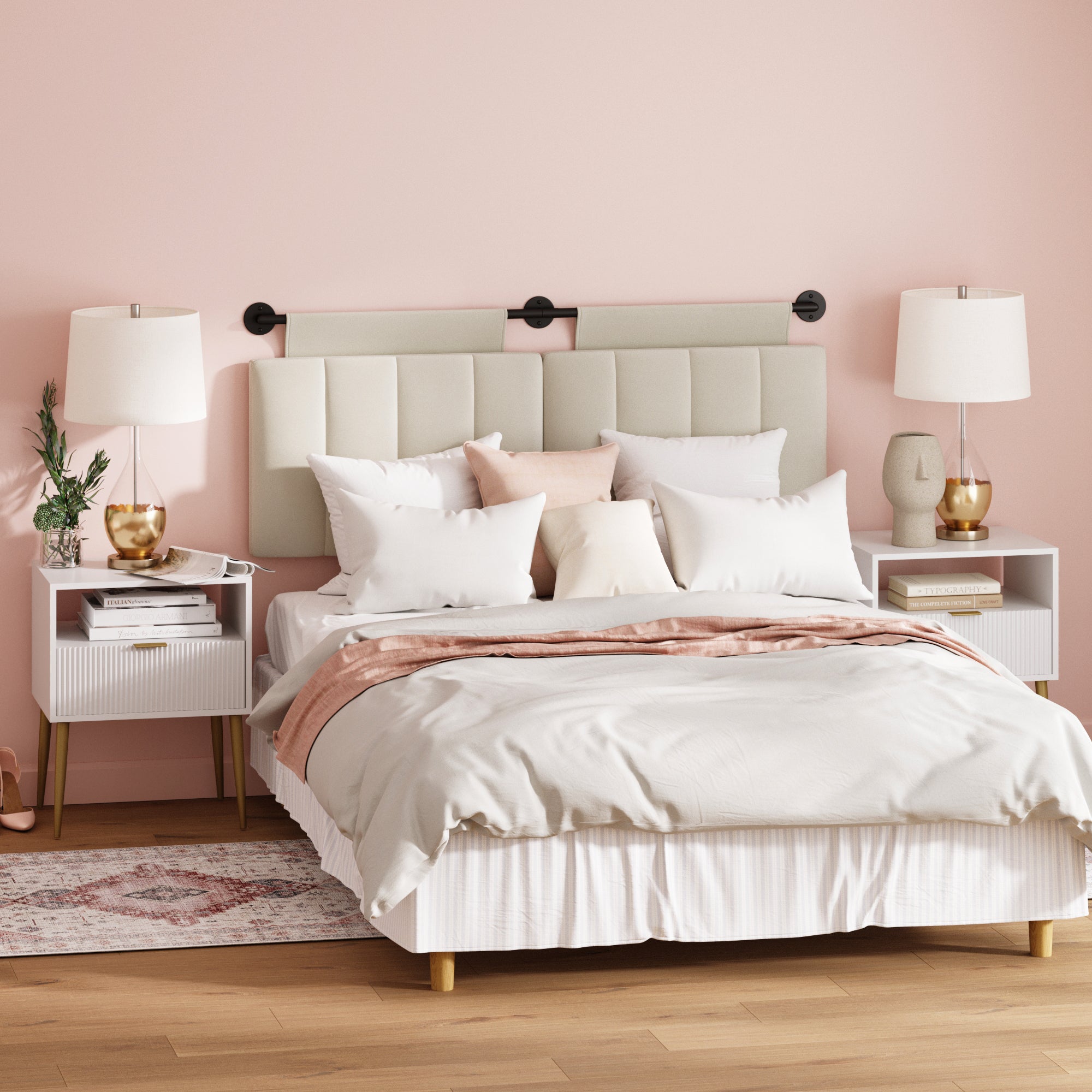 A stylish bedroom with a beige bed, white and blush bedding, two white nightstands with gold lamps, and decorative pillows, set against a soft pink wall with a modern, padded headboard.