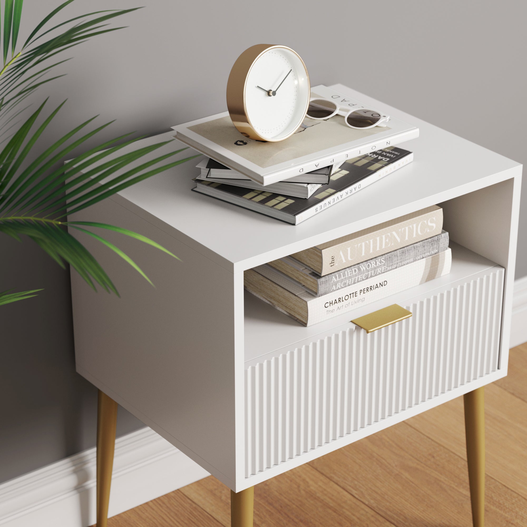 A modern white nightstand with gold legs holds books, a clock, sunglasses, and a magazine on top. A plant is partially visible to the left, and the nightstand sits on a wooden floor against a gray wall.