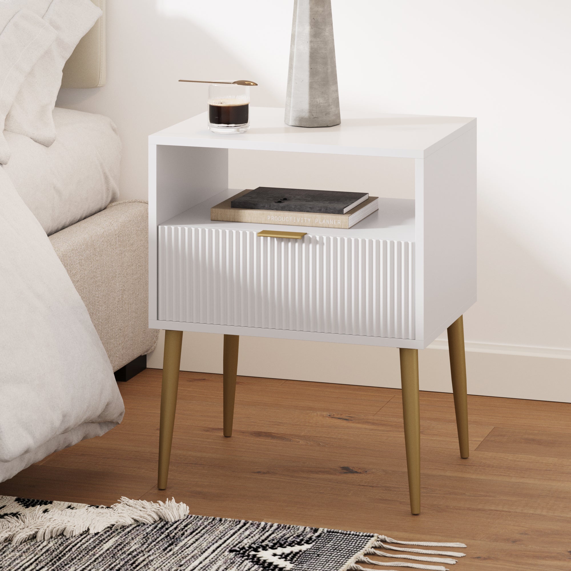 A modern white nightstand with gold legs stands beside a bed. It features one drawer with a vertical textured design and an open shelf holding a notebook. A coffee cup and a decorative vase sit on top.