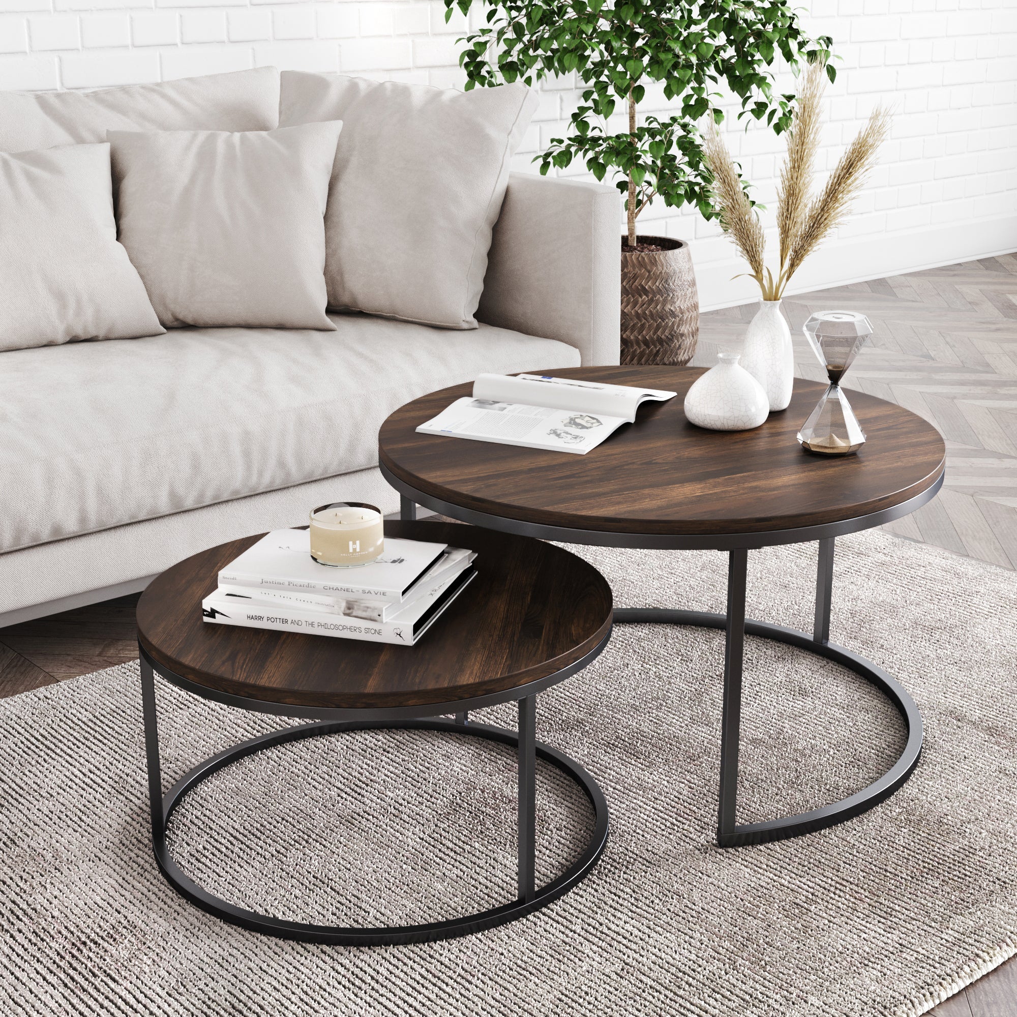 The Nathan James Round Nesting Coffee Table Set in reclaimed oak with black metal legs sits atop a textured rug in a modern living room, styled with books, a candle, vase of dried grass, and an hourglass for an industrial farmhouse look.