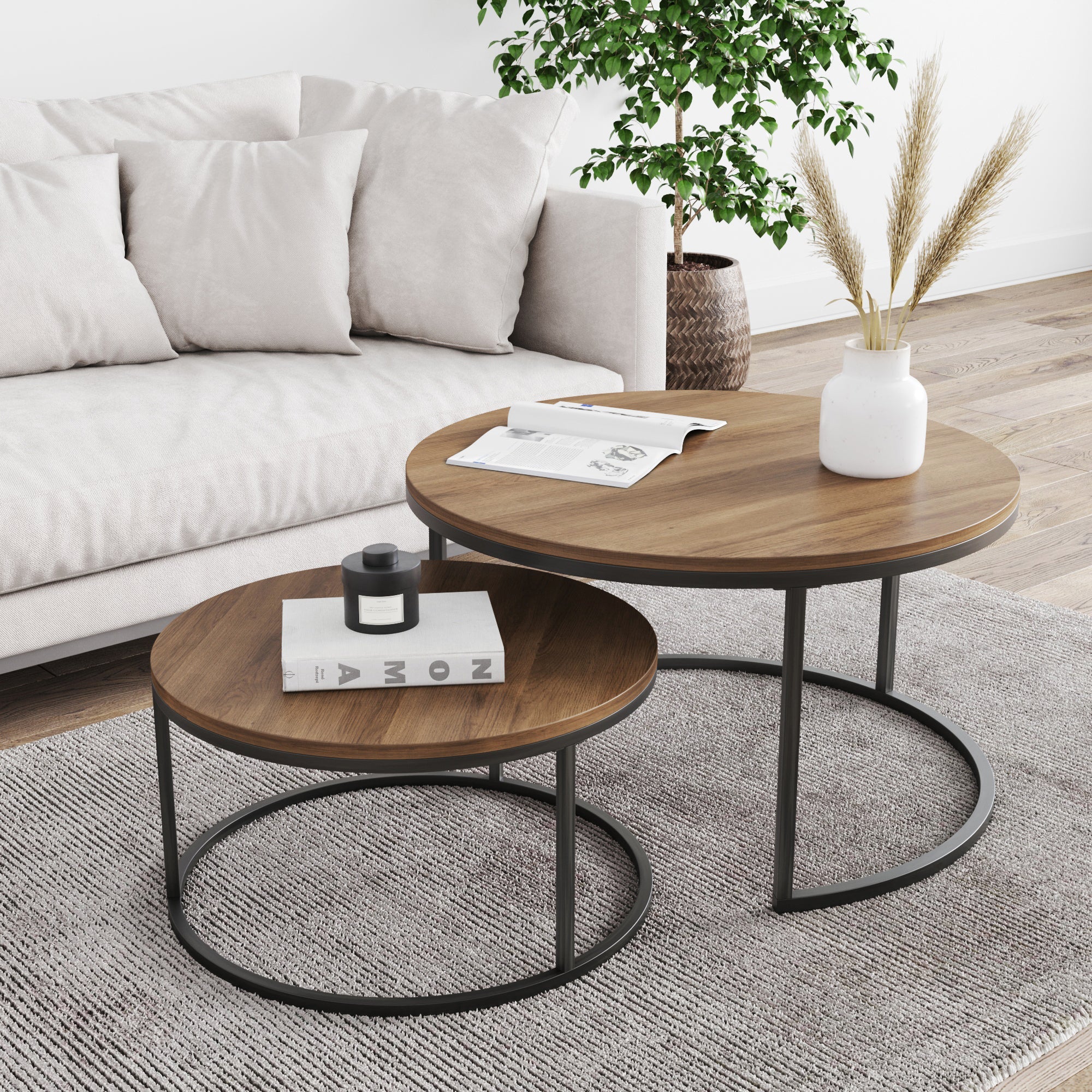 The Nathan James Round Nesting Coffee Table Set in reclaimed oak, featuring black metal frames, sits on a gray rug before a beige sofa. A white vase with dried grass and a book top the tables for an industrial farmhouse vibe.
