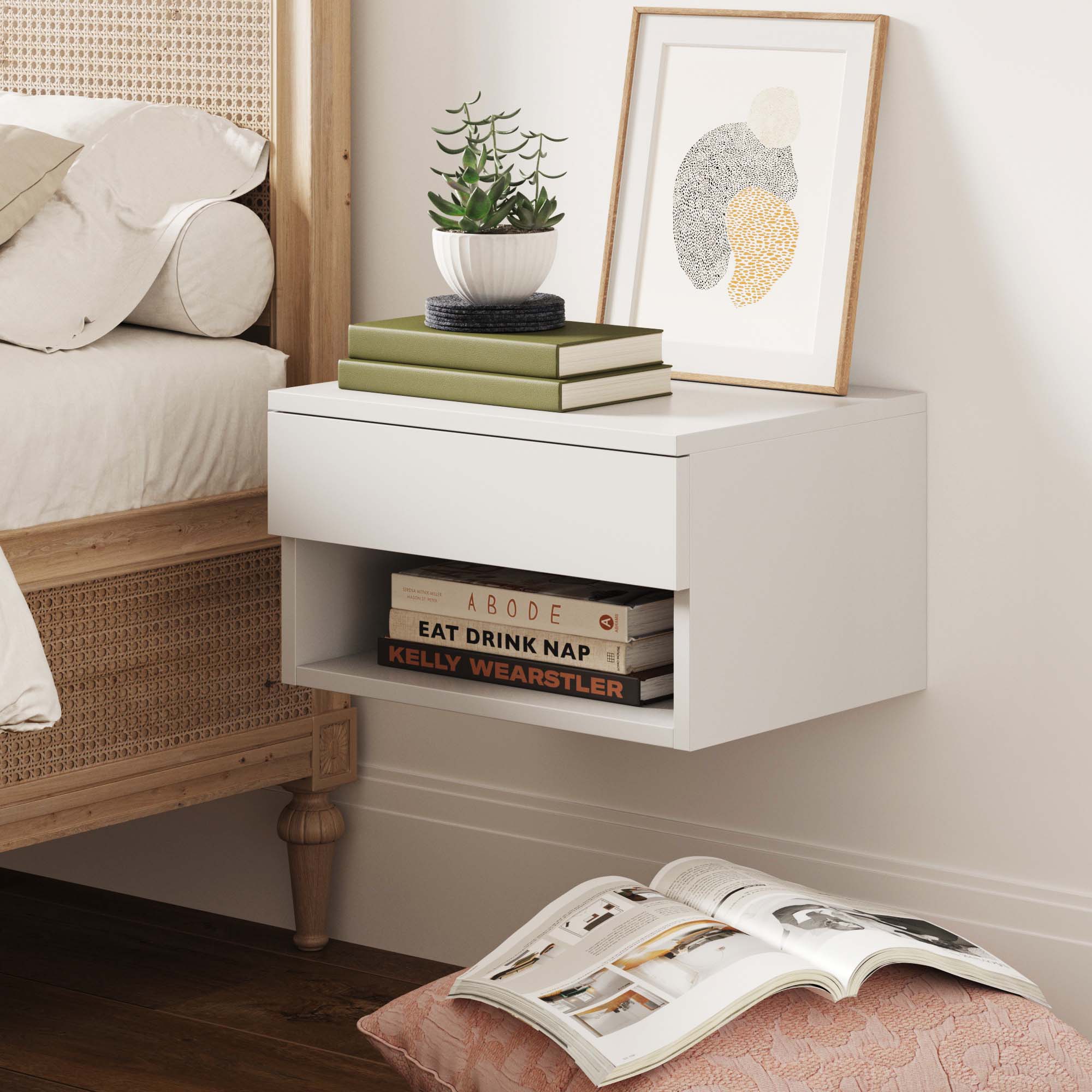 The Nathan James Mid-Century Floating Drawer Nightstand in walnut stands by the bed, topped with a potted plant, stacked books, and framed art; books fill the lower shelf while an open book lies on a pink cushion on the wooden floor.