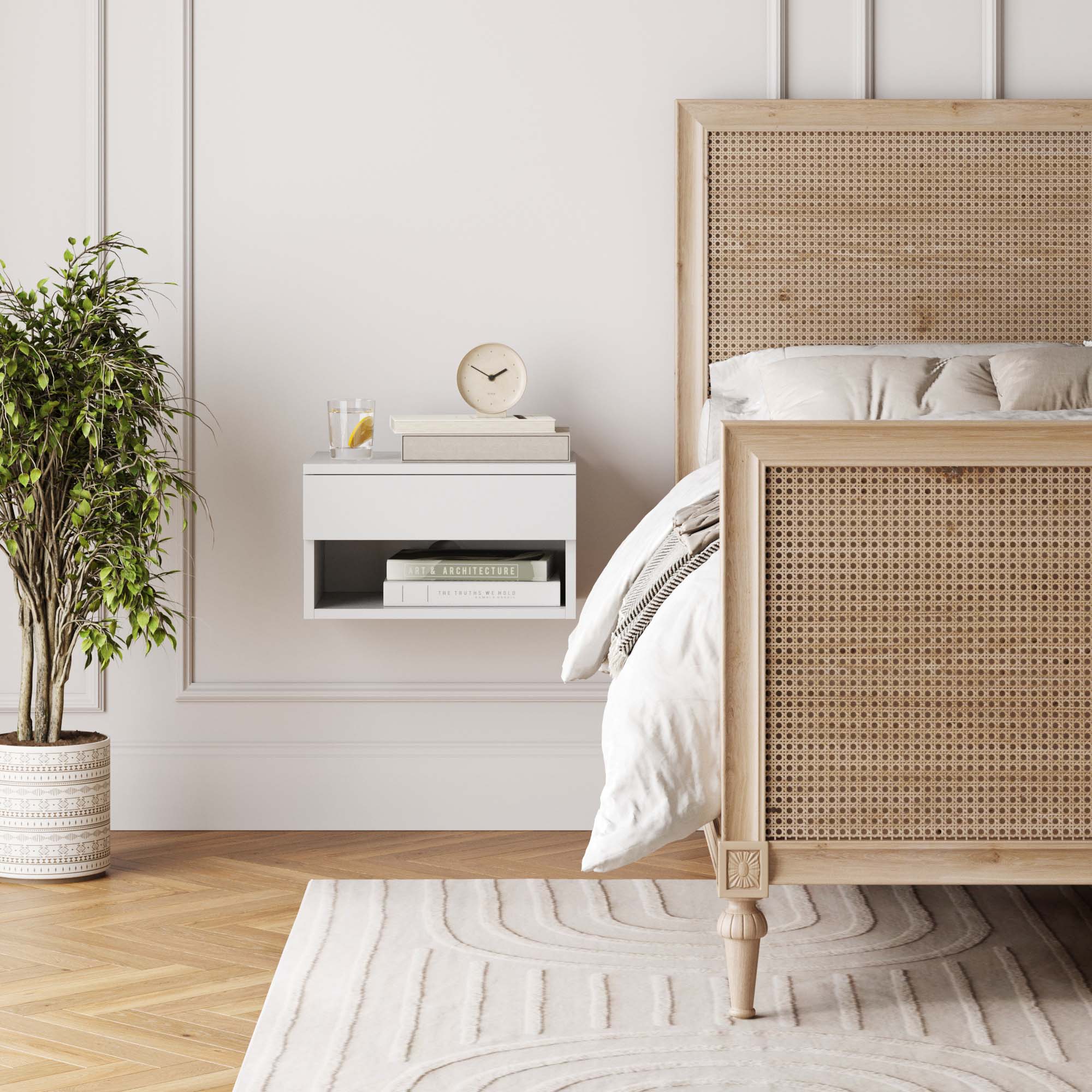 A cozy, modern bedroom with a wooden bed featuring a woven headboard, a white floating nightstand with books, a clock, and a glass of water, a large potted plant, and a patterned rug on a wooden floor.