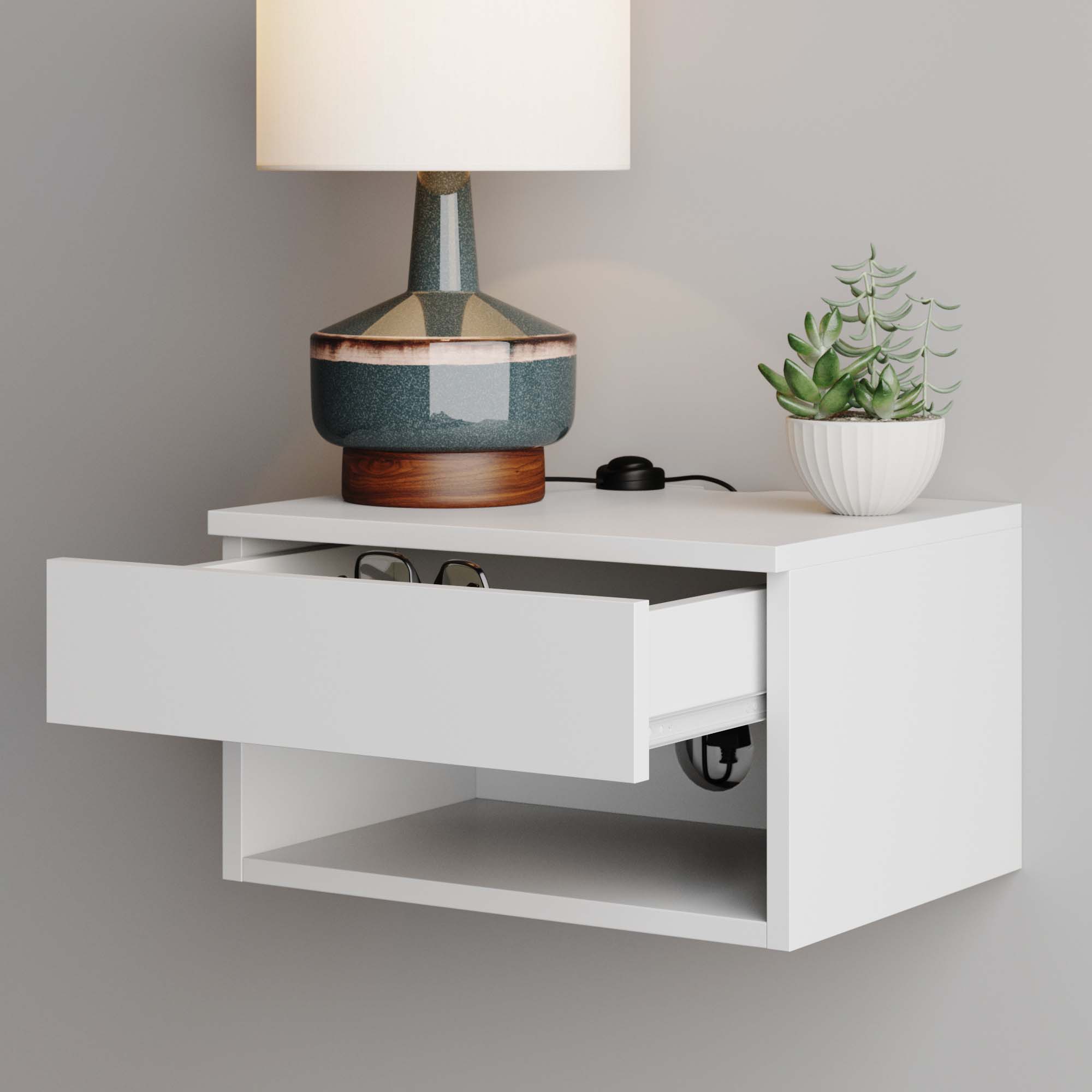 A white wall-mounted nightstand with an open drawer holding eyeglasses, a lamp with a dark and light ceramic base, a small potted succulent, and a black cord on top, against a light gray wall.