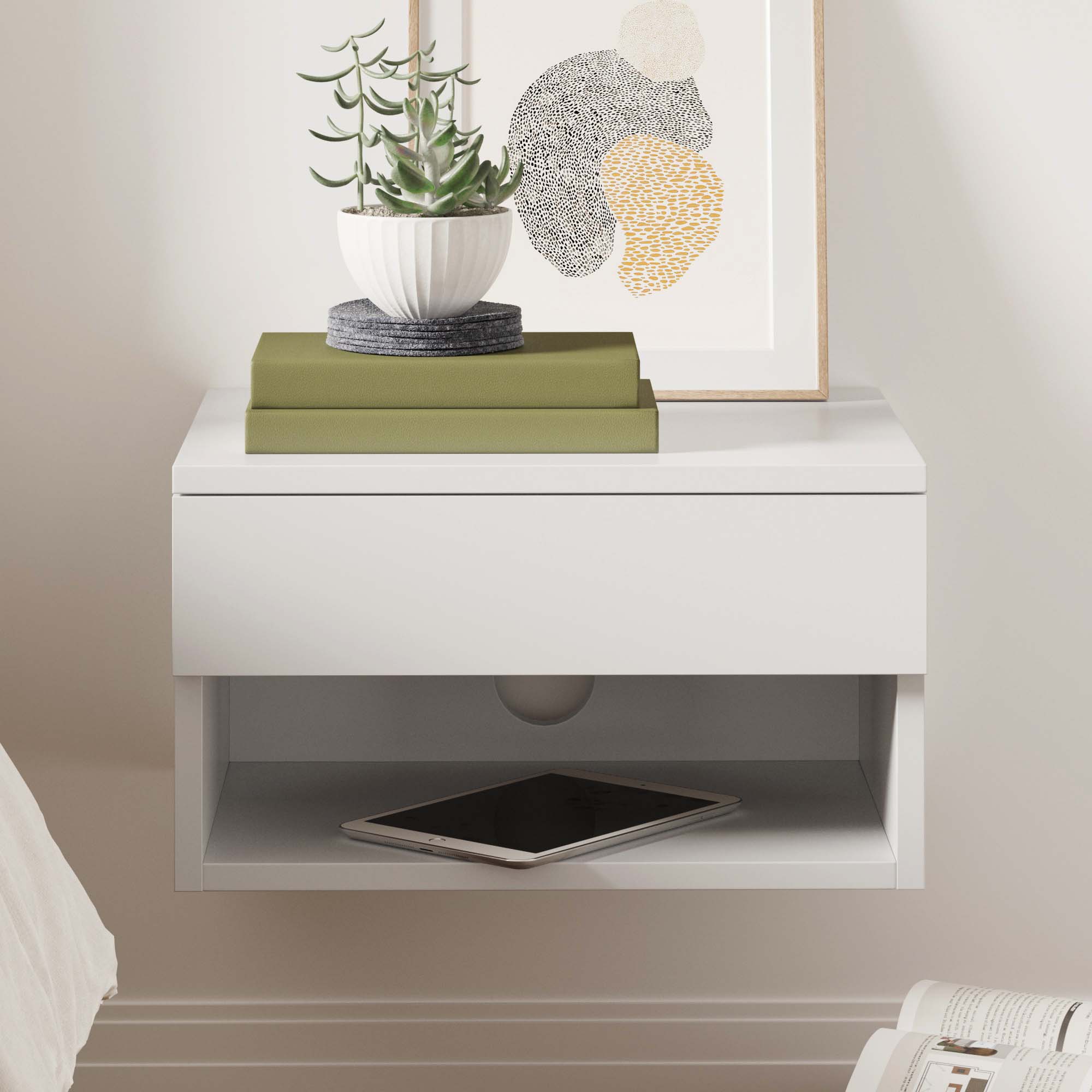 A white floating nightstand with a drawer holds two green books, a potted succulent, and framed abstract art. An electronic tablet rests on the open shelf below.