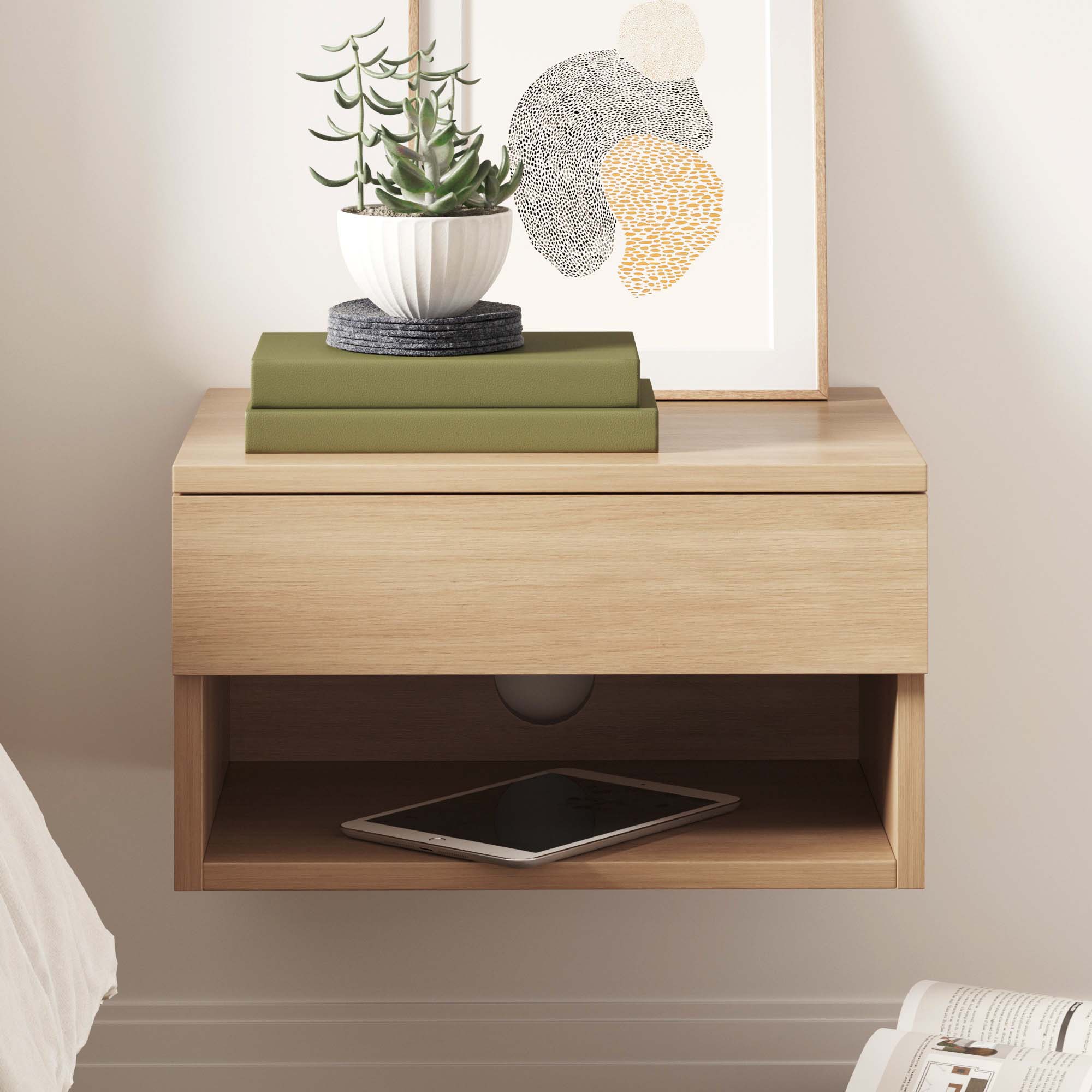 A modern wooden floating nightstand with a closed drawer, an open shelf holding a tablet, and a top surface with stacked green books, a potted succulent, and a framed abstract art print.