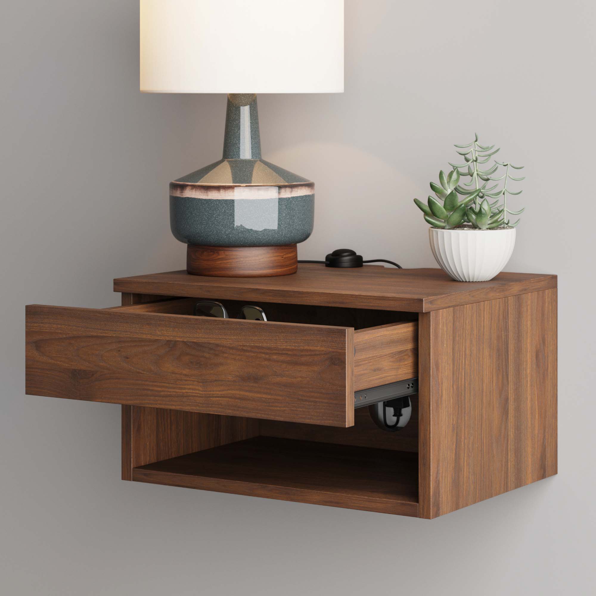 A wooden floating nightstand with an open drawer, a shelf underneath, a lamp, and a potted succulent on top. Glasses are visible inside the drawer. The nightstand is mounted on a light-colored wall.