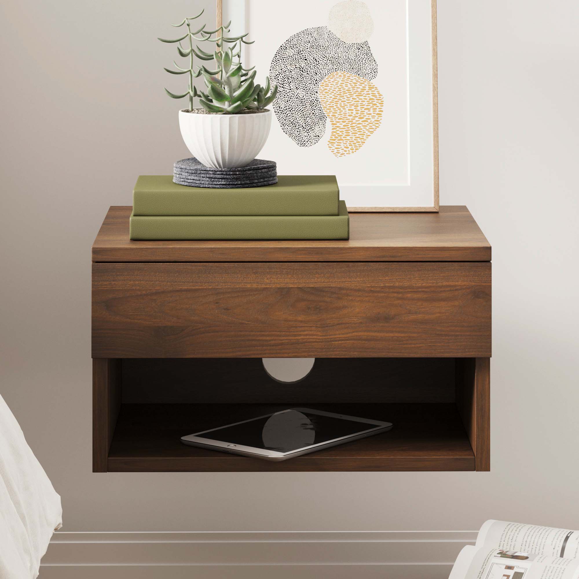 A wooden floating nightstand with a potted succulent, two stacked green books, and framed abstract art on top. An open shelf underneath holds a tablet. The nightstand is mounted to a light-colored wall.
