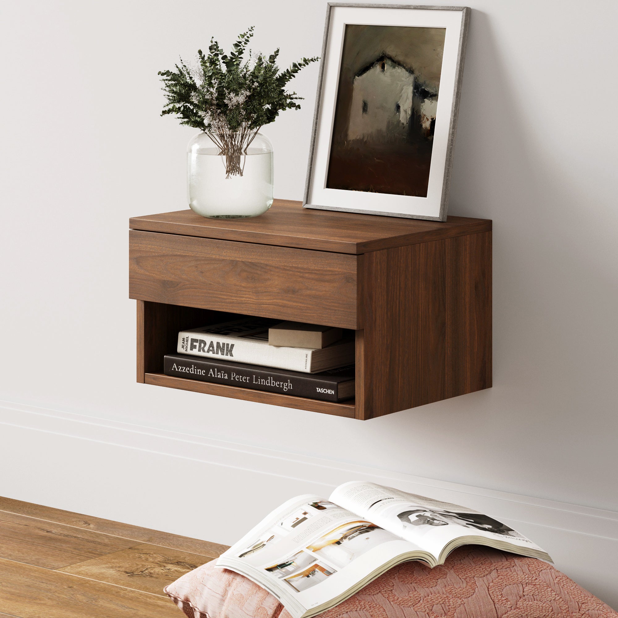 A floating wooden nightstand with a drawer, a vase of greenery, framed artwork, and books on the shelf, mounted on a white wall above a wooden floor and a pink patterned cushion with an open magazine.