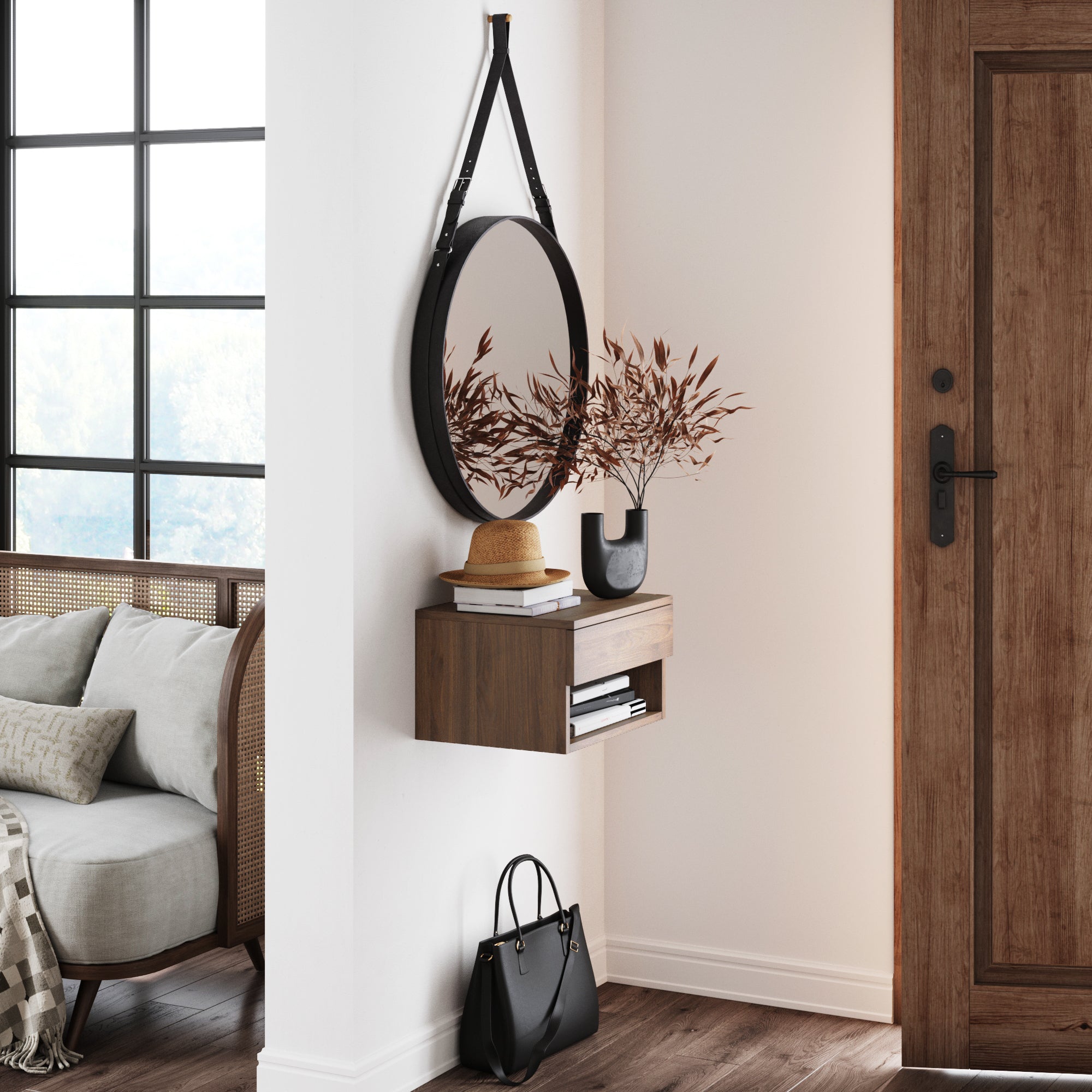 A modern entryway with a round mirror, a floating wooden shelf holding a vase of brown leaves and two books, a black handbag on the floor, and a brown door nearby. A sofa and large window are visible to the left.