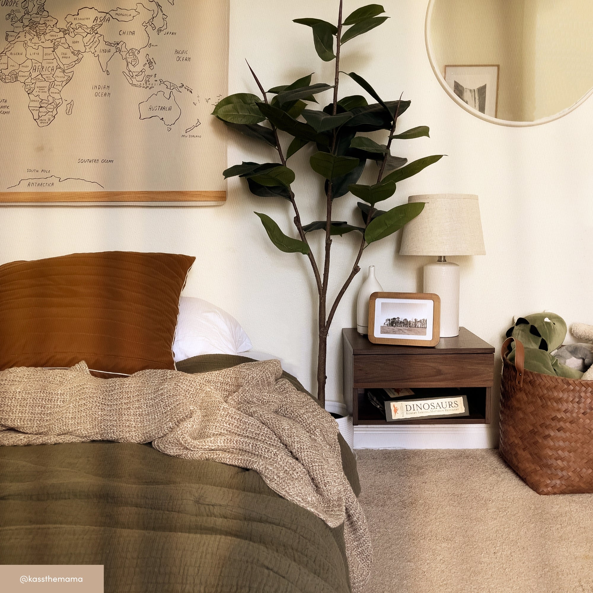 A cozy bedroom corner with an olive green bedspread, rust pillow, beige knit blanket, a world map on the wall, a tall leafy plant, round mirror, nightstand with a lamp and photo, and a basket with plush toys.
