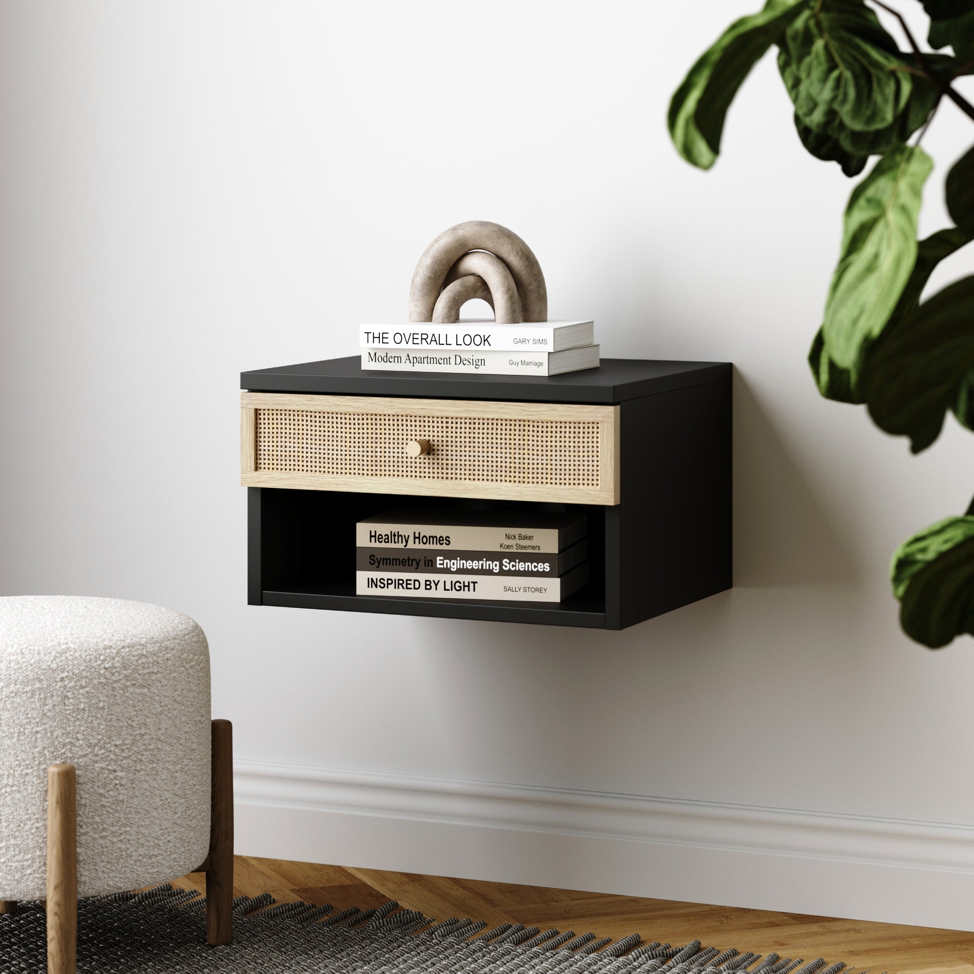 The Nathan James Rattan & Wood Floating Nightstand in black features a wicker drawer and modern design above a wood floor. Nearby, a green plant and round upholstered stool complement the white wall for a stylish bedside look.