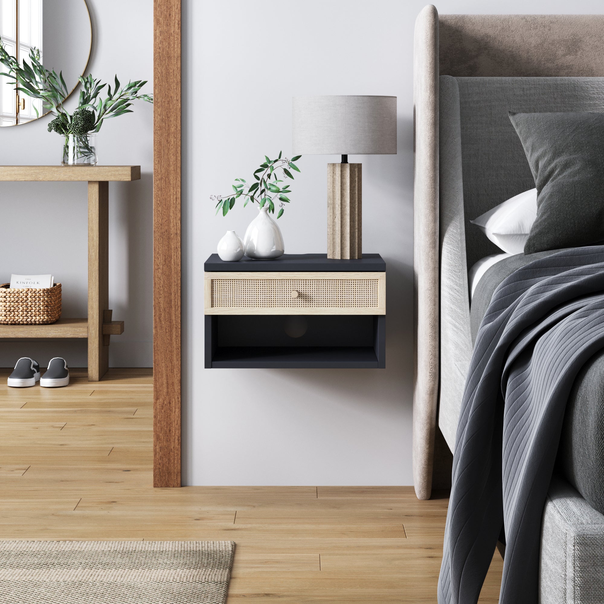 A modern bedroom features a floating nightstand with a drawer, a table lamp, and vases with greenery next to a gray bed. Wooden floors, a rug, and a console table with baskets and decor are visible in the background.