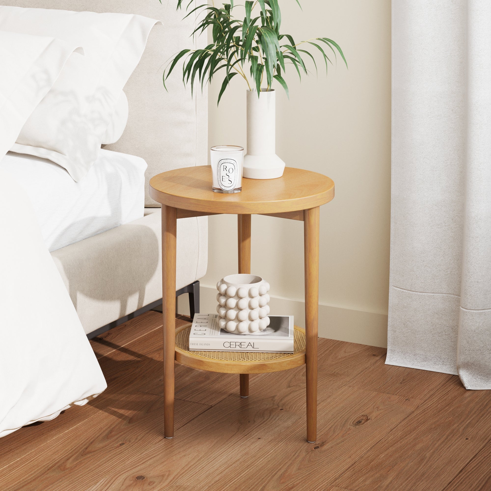 The Nathan James Wood & Rattan Round End Table in Warm Pine holds a vase with greenery and a candle on top; books and a decorative white bubble candle rest on the lower shelf, complementing the beige bed and white curtains.