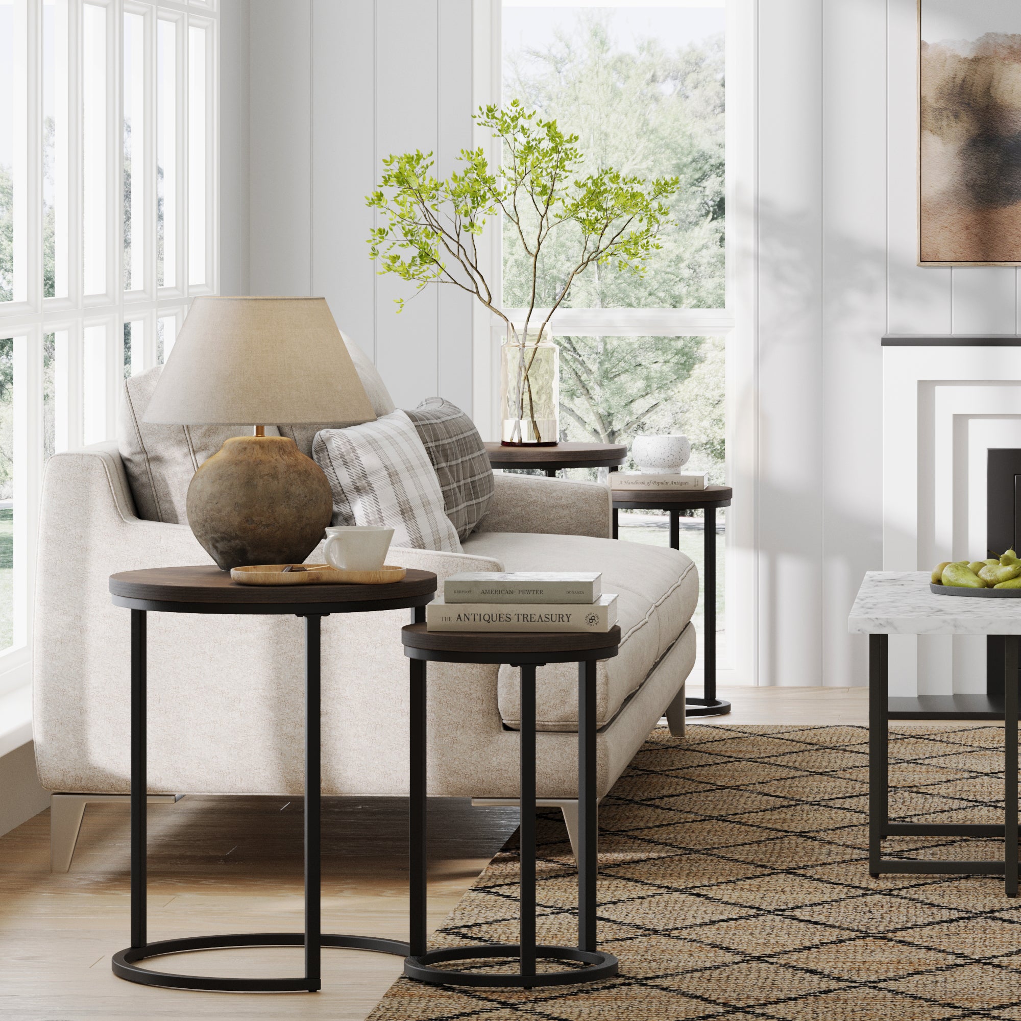 A cozy living room with a beige sofa, plaid pillows, and Nathan James Nesting Round End Table Set in Black/Nutmeg holding a rustic lamp, books, and a vase with greenery. Large windows fill the modern space with natural light.