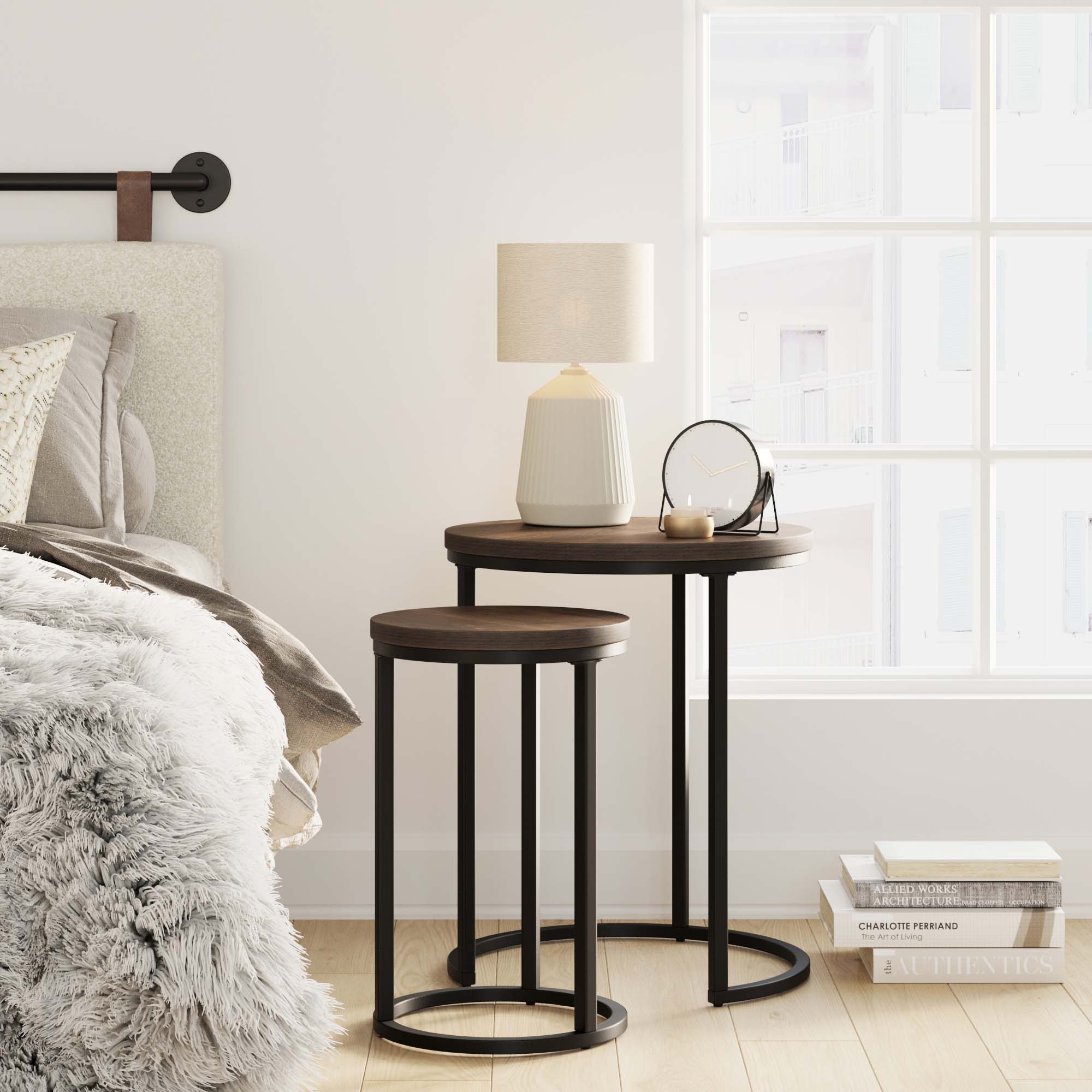 A cozy bedroom corner with two round nesting tables, a white ceramic lamp, a small mirror, and books. The tables sit beside a bed with a furry gray blanket, near a window letting in natural light.