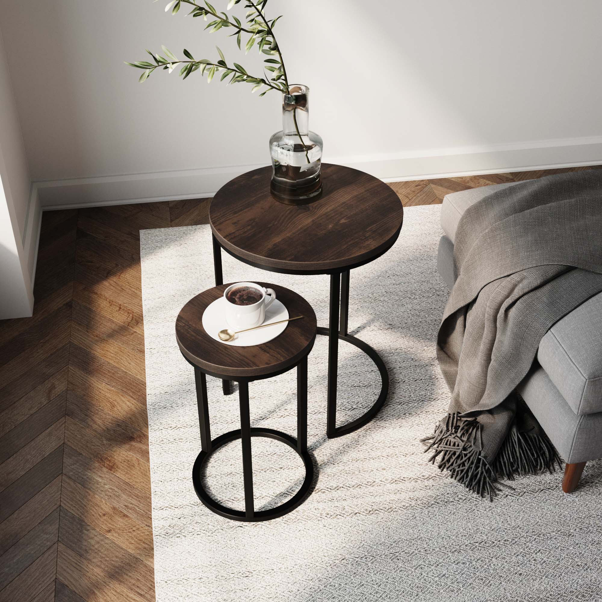 The Nathan James Nesting Round End Table Set in nutmeg with black metal legs sits on a light rug by a gray sofa with a fringe blanket. A coffee cup is on the smaller table, and a glass vase with greenery decorates the larger one.