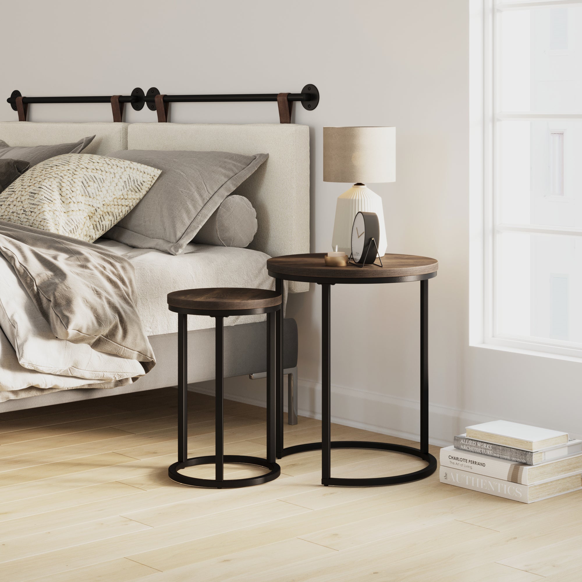 A modern bedroom with a beige bed, gray bedding, and two round nesting tables holding a lamp, clock, and sunglasses. Sunlight streams through a large window, and books are stacked on the wooden floor.