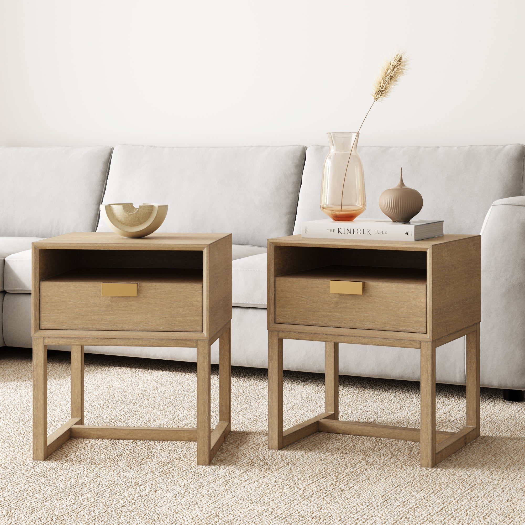 Two Nathan James Wood Modern Farmhouse Nightstands (Set of 2) are on a beige carpet by a light gray sofa—one holds a decorative bowl, the other displays a clear vase with dried grass and a small brown vase on top of a book.