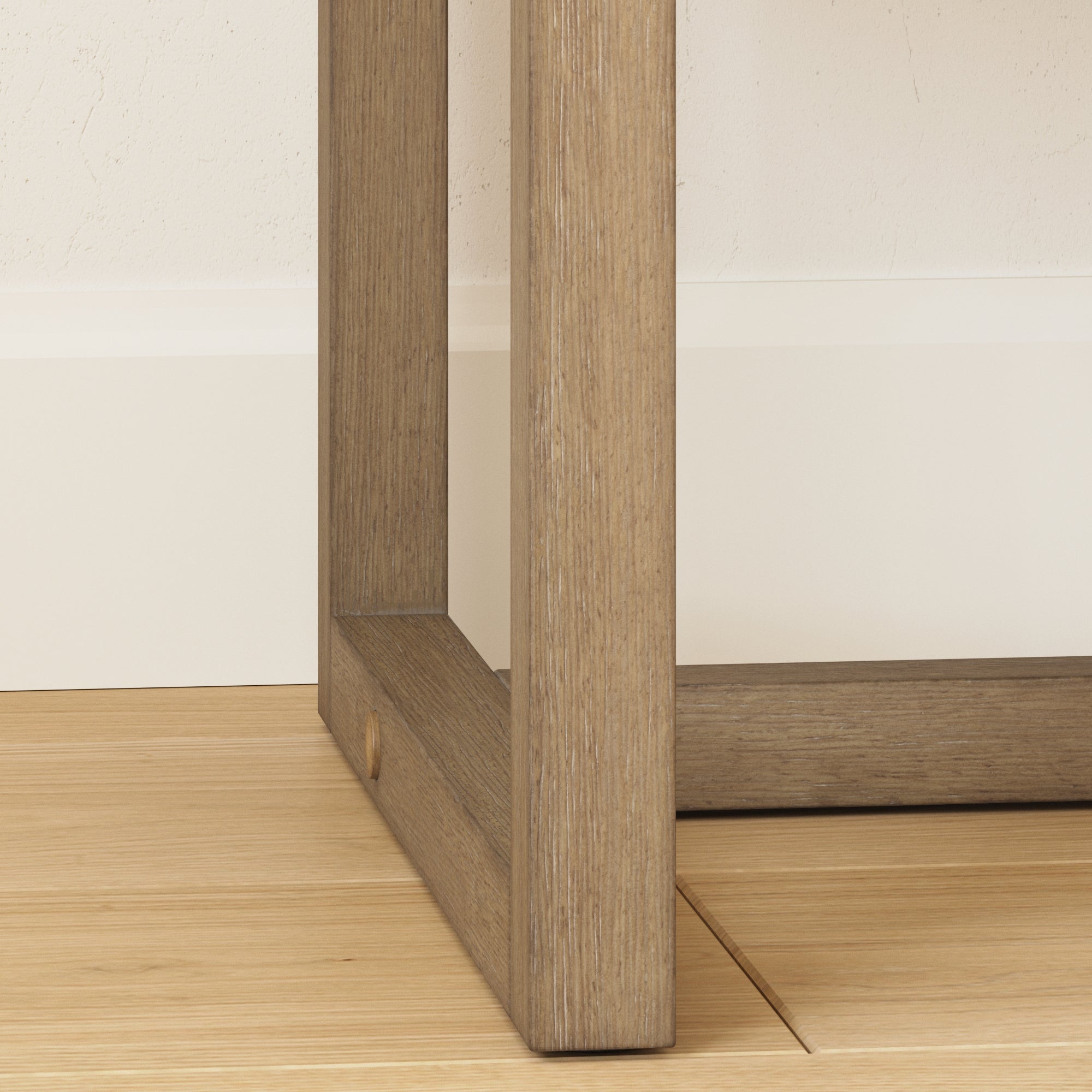 Close-up of the wooden leg and base of a table or furniture piece on a light wood floor, with a white baseboard and textured white wall in the background.