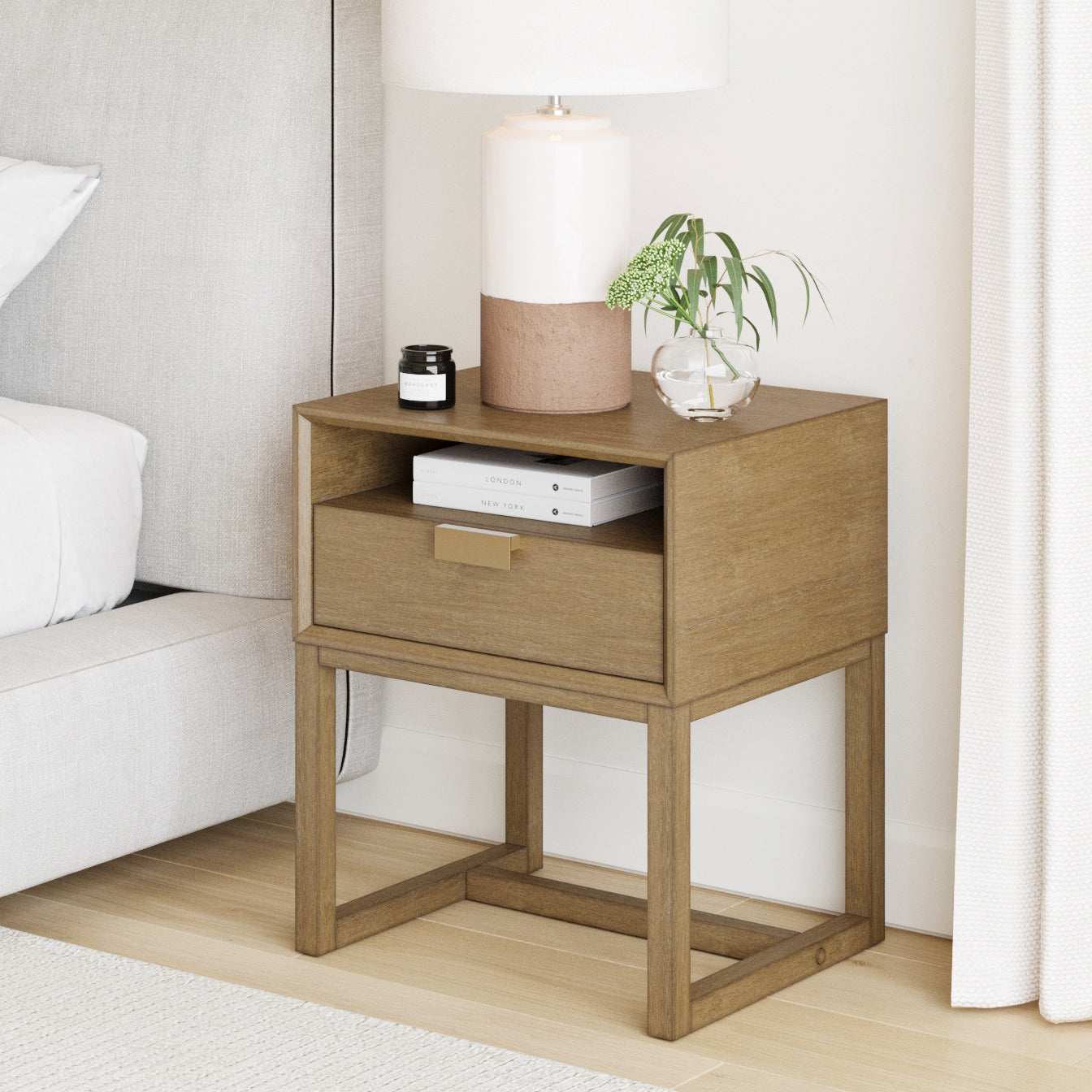 A wooden nightstand with an open shelf and drawer holds a lamp, a small plant in a glass vase, two stacked books, and a small black jar. The stand sits beside a light-colored bed and curtains.