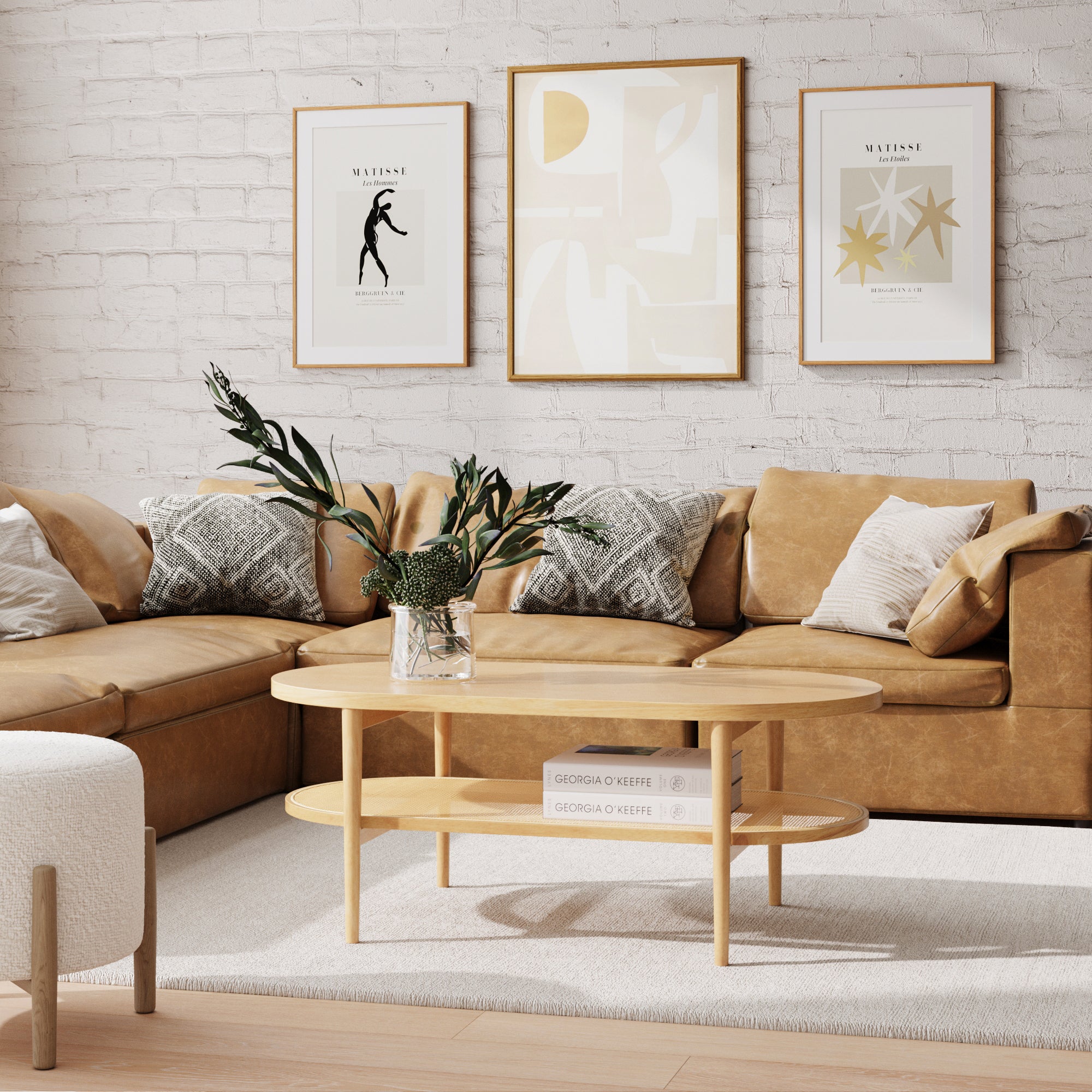 A cozy living room features a tan leather sectional sofa with patterned pillows, a round wooden coffee table with a plant and books, light rug, and three framed abstract artworks on a white brick wall.