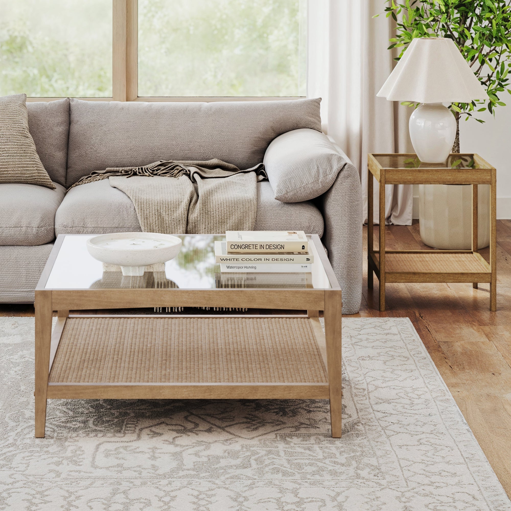 A modern living room features Nathan Jamess Glass Coffee Table & End Table Set with Rattan Shelf, styled with books, a bowl, a lamp, and a plant atop boho tables, plus a patterned rug and large windows—ideal for small space furniture enthusiasts.