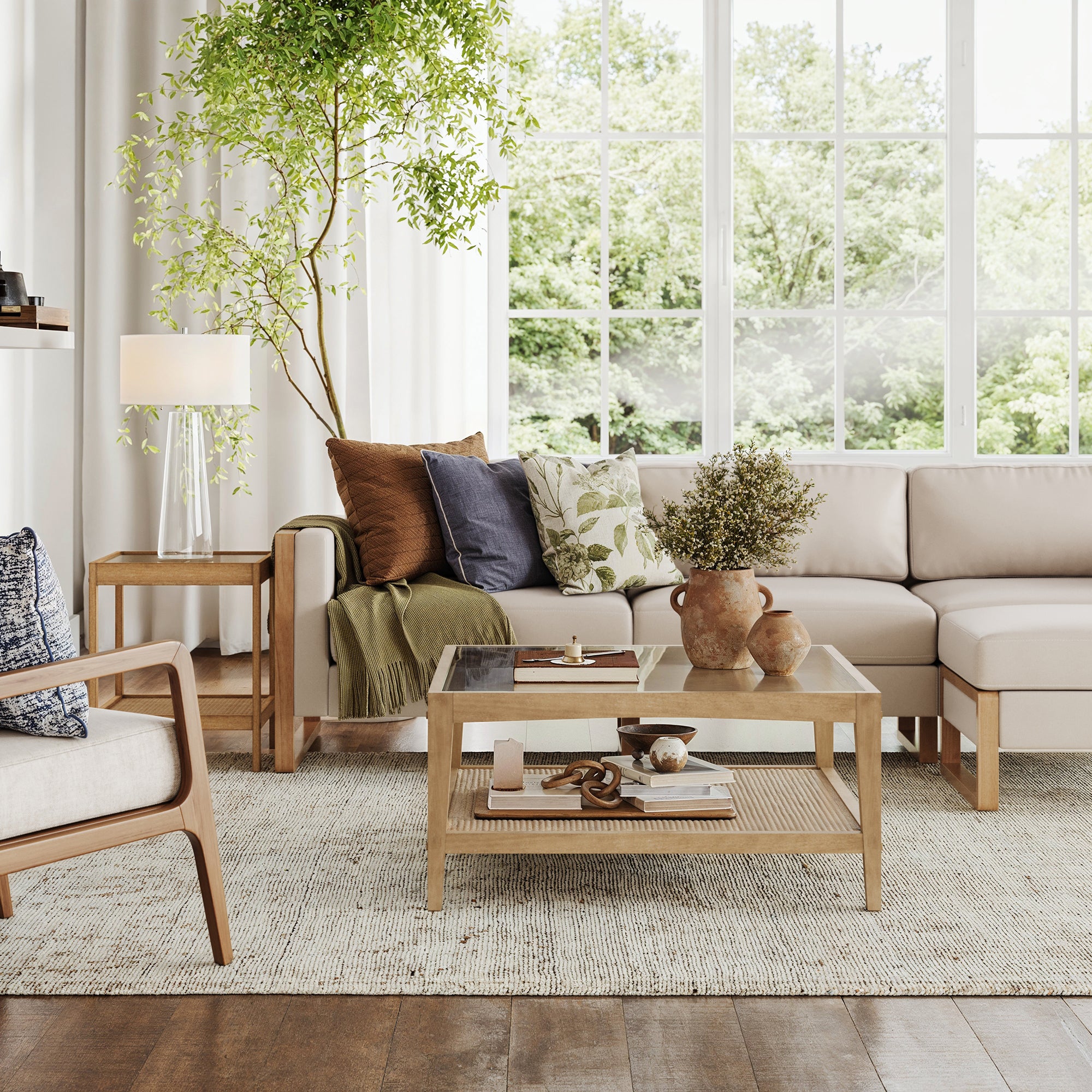 A bright living room with a beige sectional, small space furniture, an armchair, a boho side table with lamp, large windows, tall potted plant, and Nathan James Glass Coffee Table & End Table Set with Rattan Shelf; green trees are visible outside.