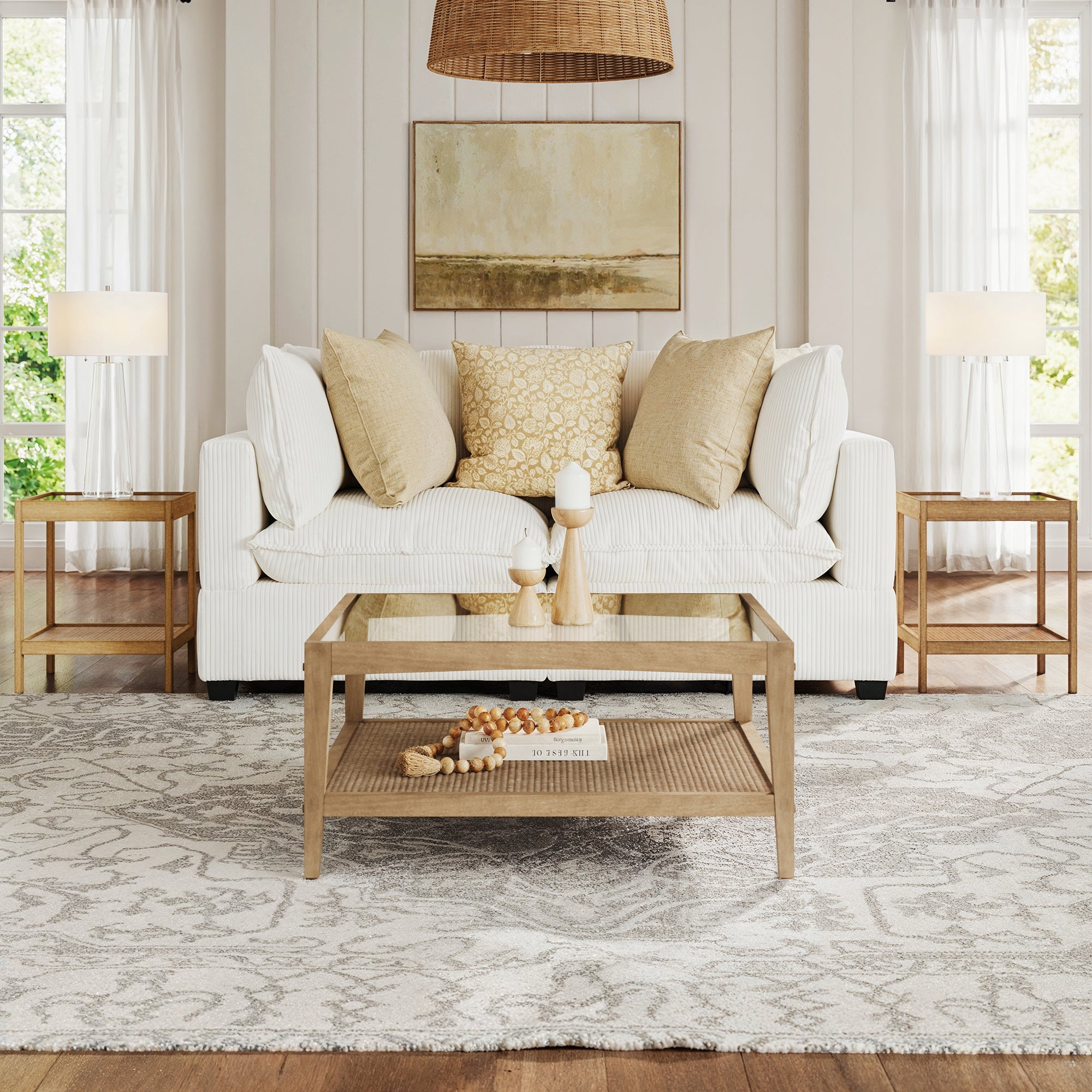 A cozy living room features the Nathan James Glass 3-Piece Coffee Table Set with a rattan shelf, a white sofa, beige and patterned pillows. Two side tables with lamps and neutral decor complete this bright, small-space furniture setup.