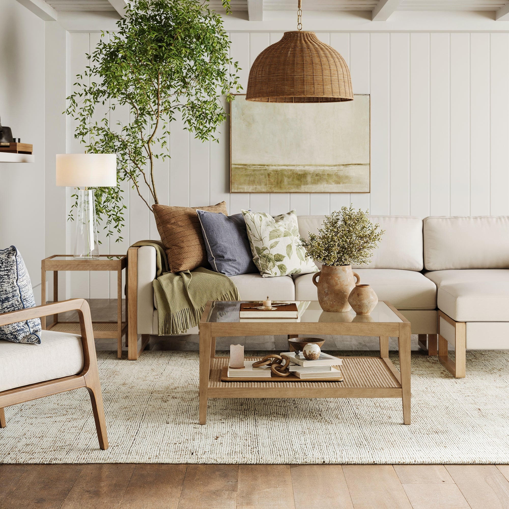 A modern living room featuring a beige sectional, wooden armchair, Nathan James Square Glass & Rattan Coffee Table, large plant, floor lamp, textured rug, and woven pendant light creates a cozy, natural vibe.