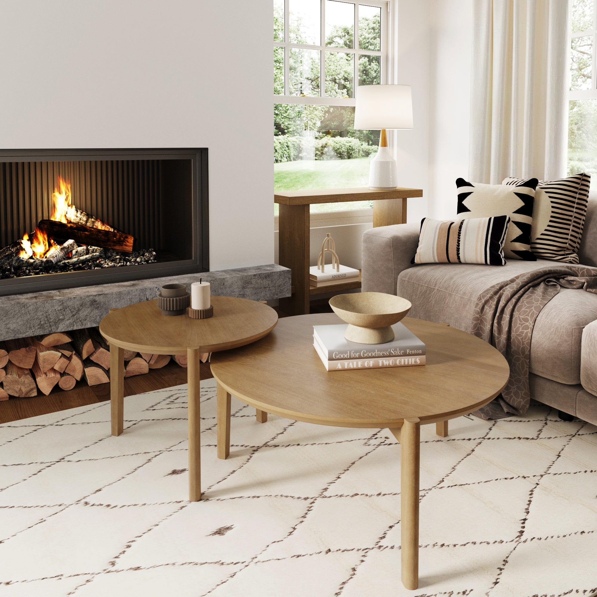A cozy living room with a lit fireplace, beige sofa with patterned cushions, a Nathan James Mid-Century Round Nesting Coffee Table Set in light brown, lamp, decor accents, stacked firewood below the fireplace, and a window overlooking greenery.